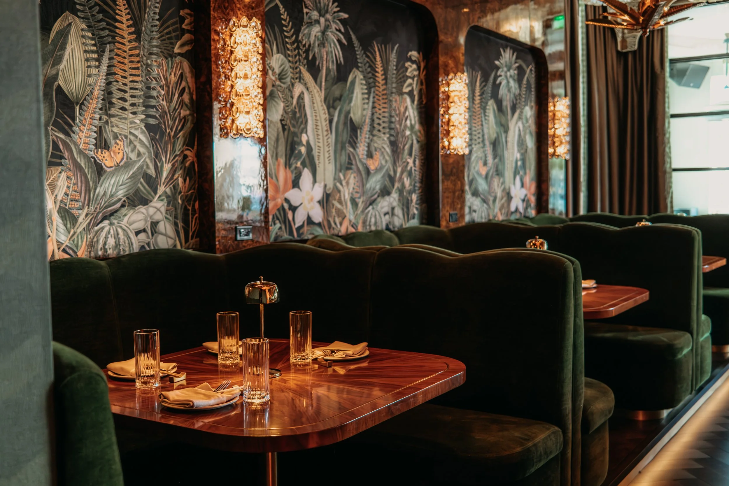 Upscale restaurant with dark green upholstered booths, wooden tables set with glassware and napkins, floral wallpaper with butterflies, and wall sconces providing warm lighting.