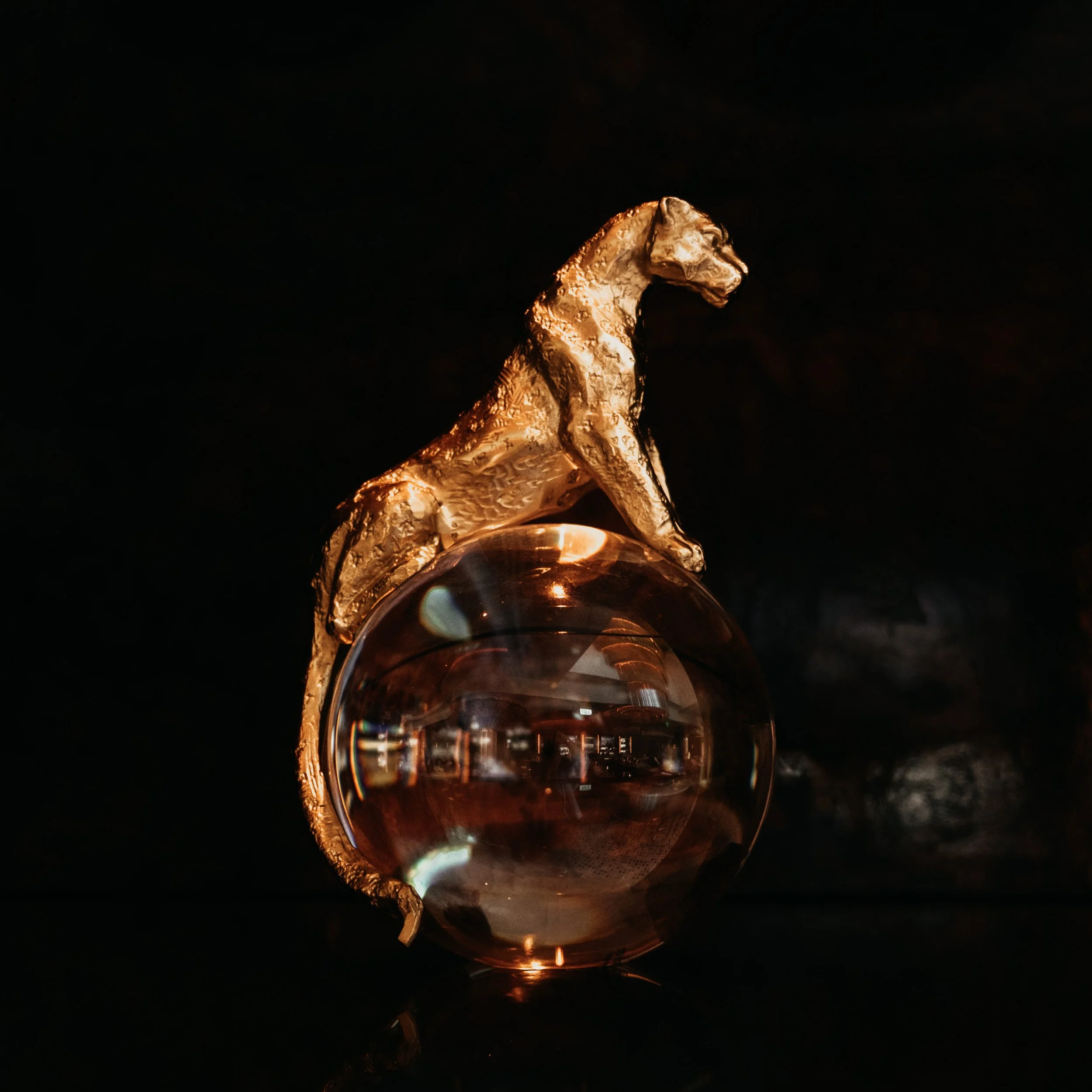 A gold-colored statue of a sitting dog perched on a reflective glass sphere, against a dark background.
