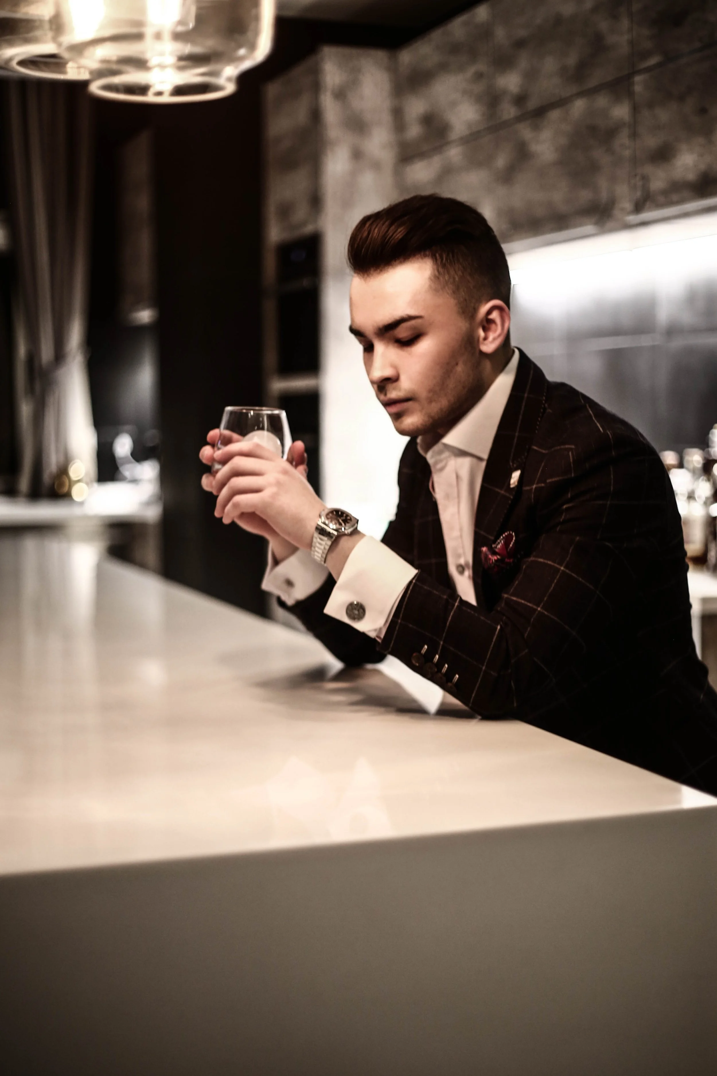 Young man in a suit sitting at a bar, holding a glass of liquor, with a focused expression.