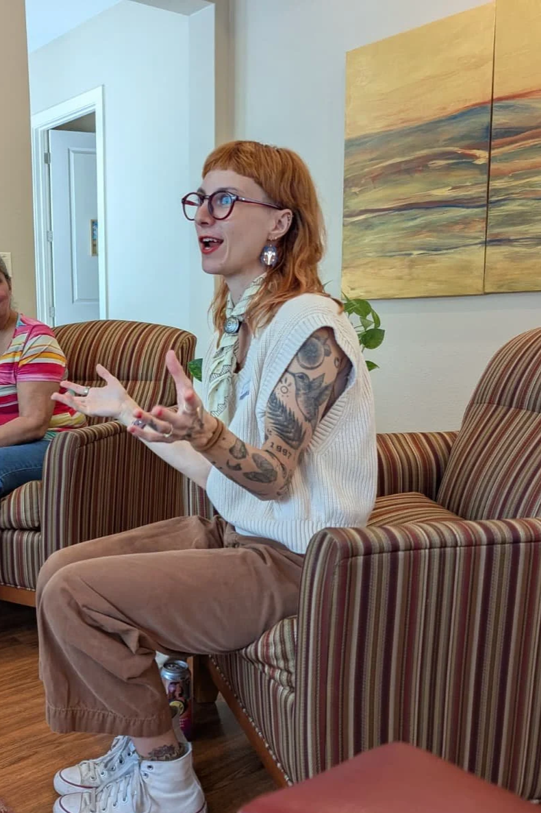 A woman with red hair, glasses, and tattoos on her arms is sitting on a striped armchair, engaging in conversation with a smile, while gesturing with her hands. She is wearing a white sleeveless top and brown pants. In the background, there is a colorful abstract painting and another person wearing a pink striped shirt.