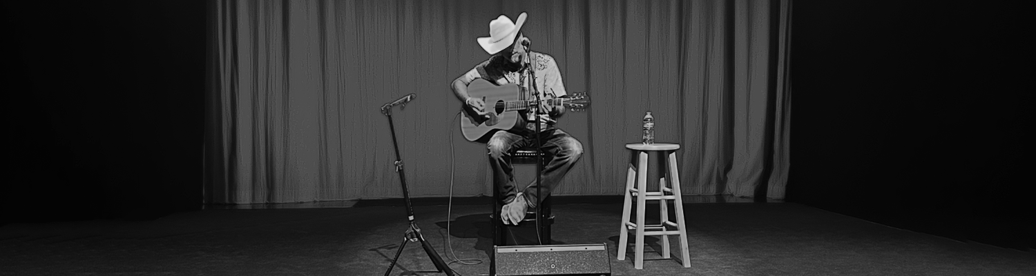A man wearing a cowboy hat and jeans playing an acoustic guitar on a stage with a curtain backdrop, a microphone stand, a small table with a water bottle, and a stool.