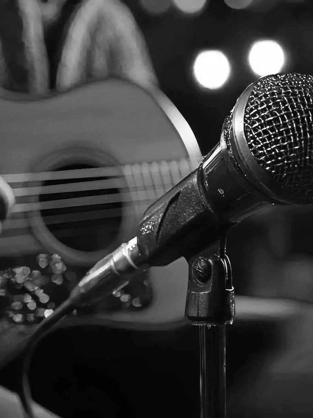 Close-up of a microphone and an acoustic guitar in a dark setting, with some blurred bright lights in the background.