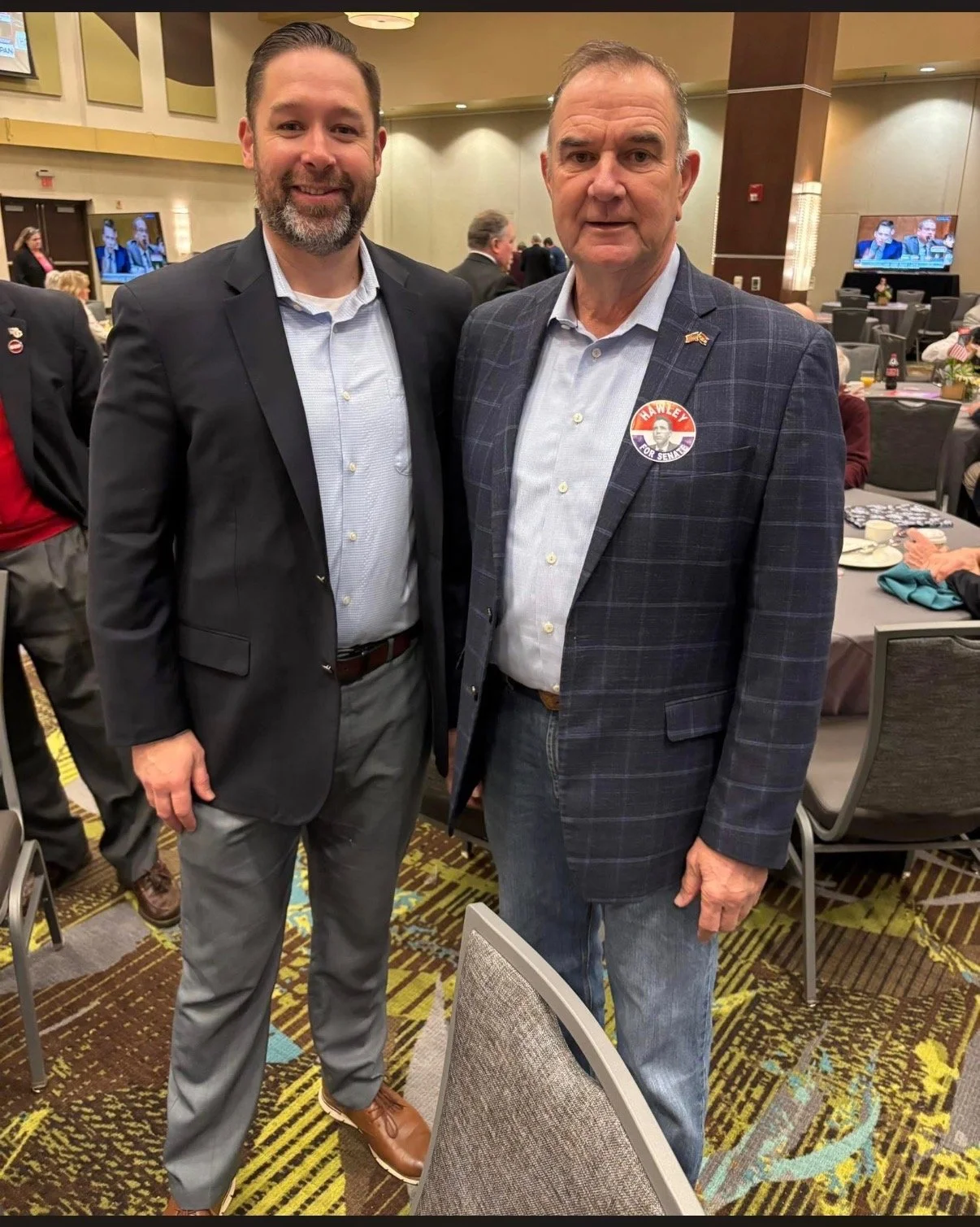 Two men posing together at a formal event, one wearing a dark suit and the other wearing a checked blazer with a campaign sticker.