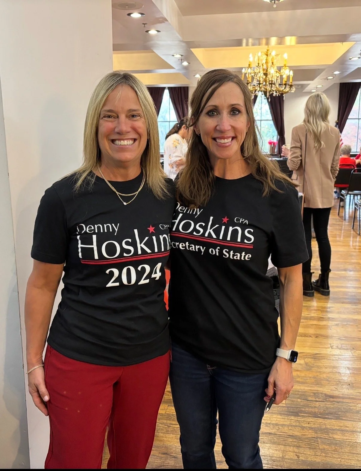 Two women smiling and wearing black T-shirts that say 'Denny Hoskins 2024' and 'Secretary of State'. They are standing in a room with a chandelier and large windows, with other people in the background.