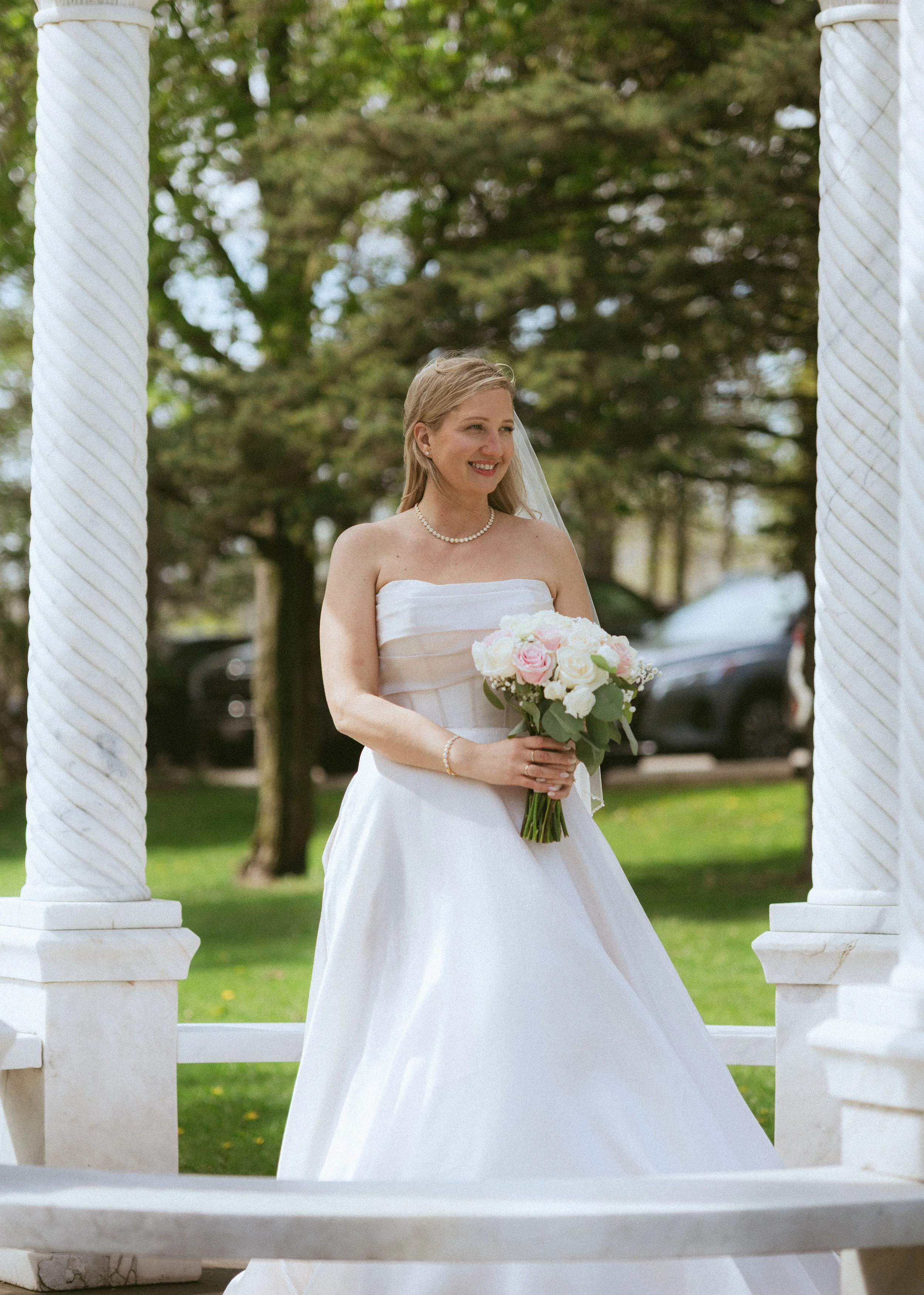 A bride in a white strapless wedding dress holding a bouquet of pink and white roses, standing outdoors between two white columns with a green, tree-filled background, smiling.
