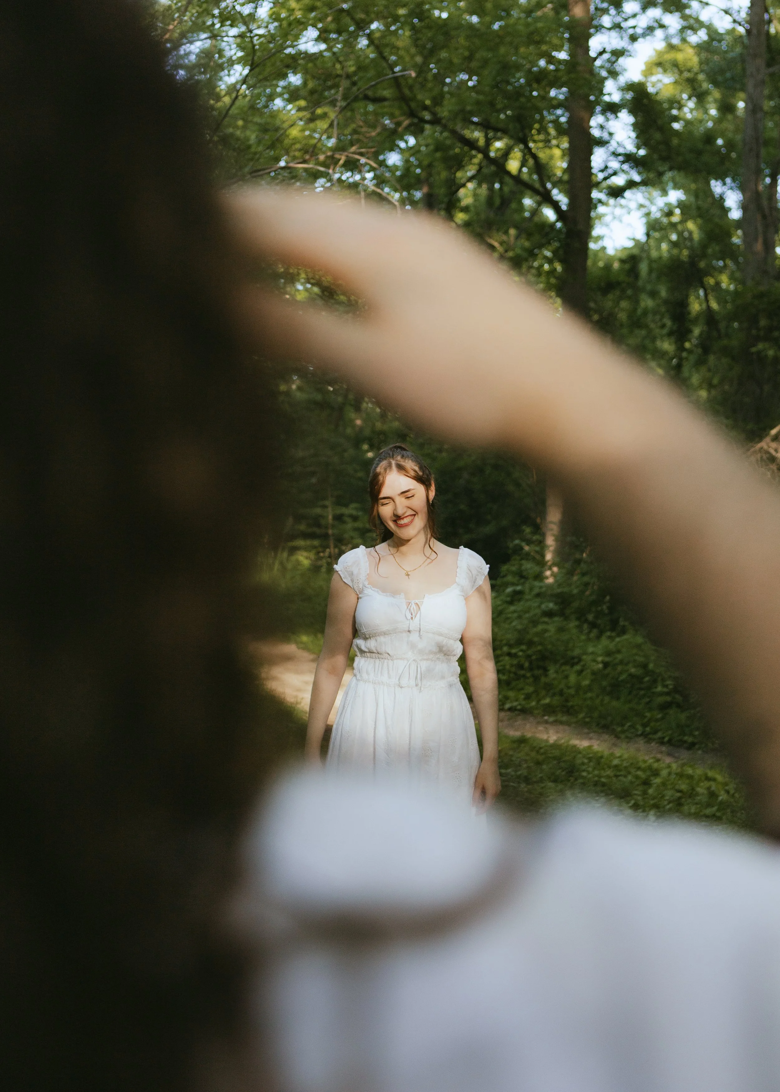 A young woman in a white dress with a smiling face, standing outdoors in a green, wooded area.