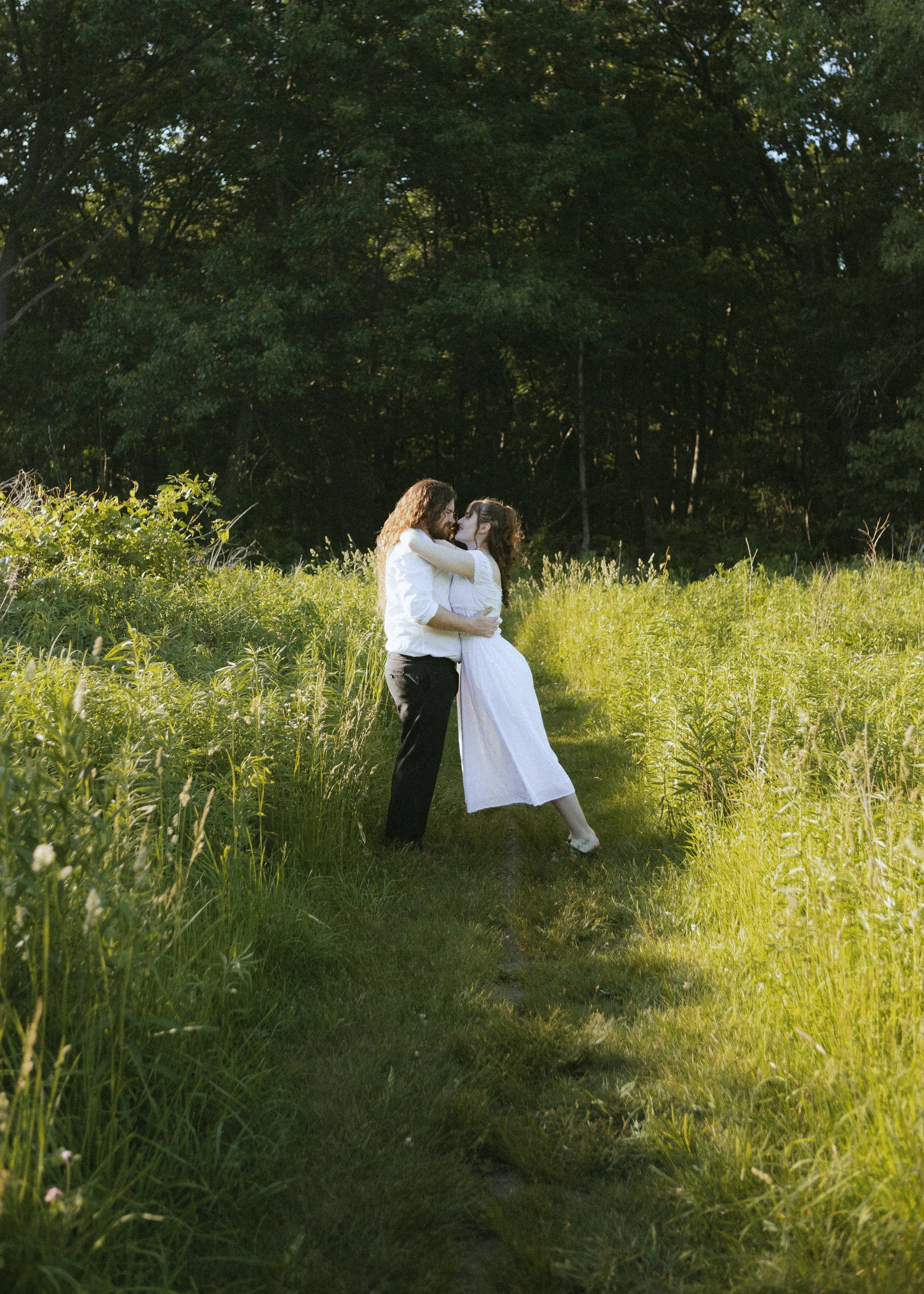 A couple embracing in a grassy field with a dark forest in the background, illuminated by sunlight.