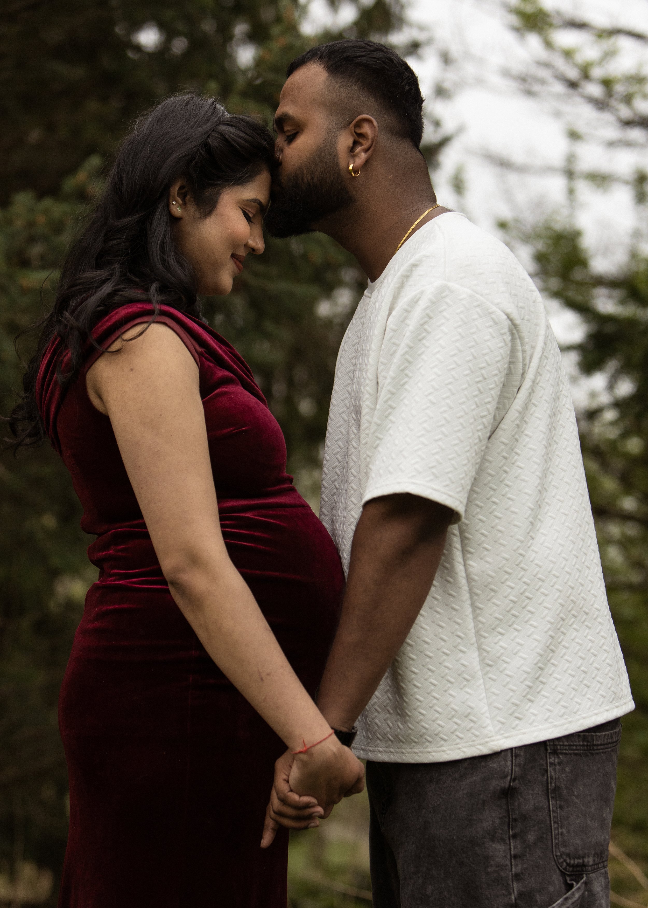 A couple holding hands outdoors, with the man kissing the woman's forehead. The woman is wearing a red velvet dress, and the man is wearing a white textured shirt. They are standing close together in a natural setting with trees in the background.
