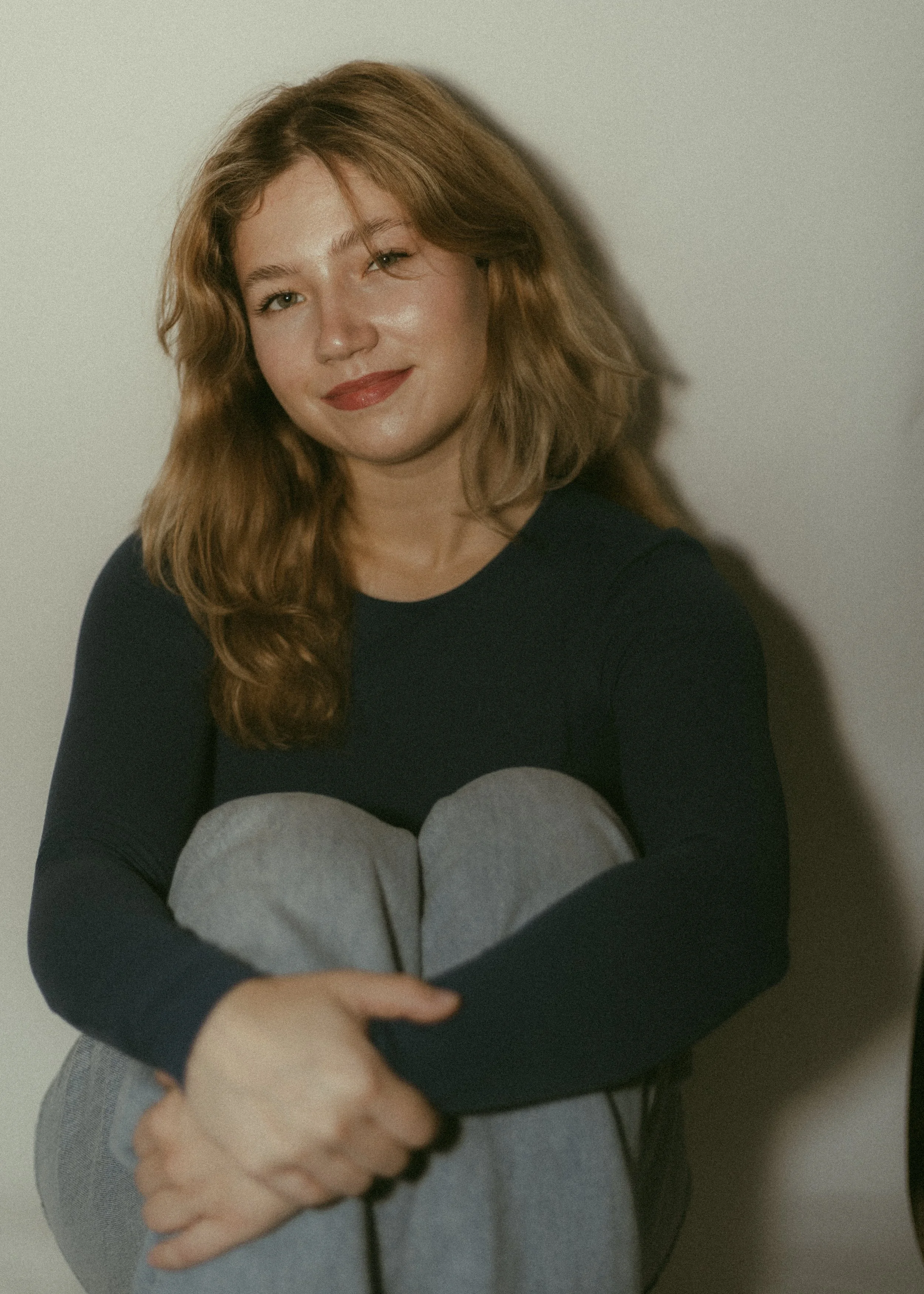 A woman with red hair and green eyes, smiling, sitting against a plain background, wearing a black top and gray sweatpants, with her knees pulled close to her chest and her arms wrapped around her knees.