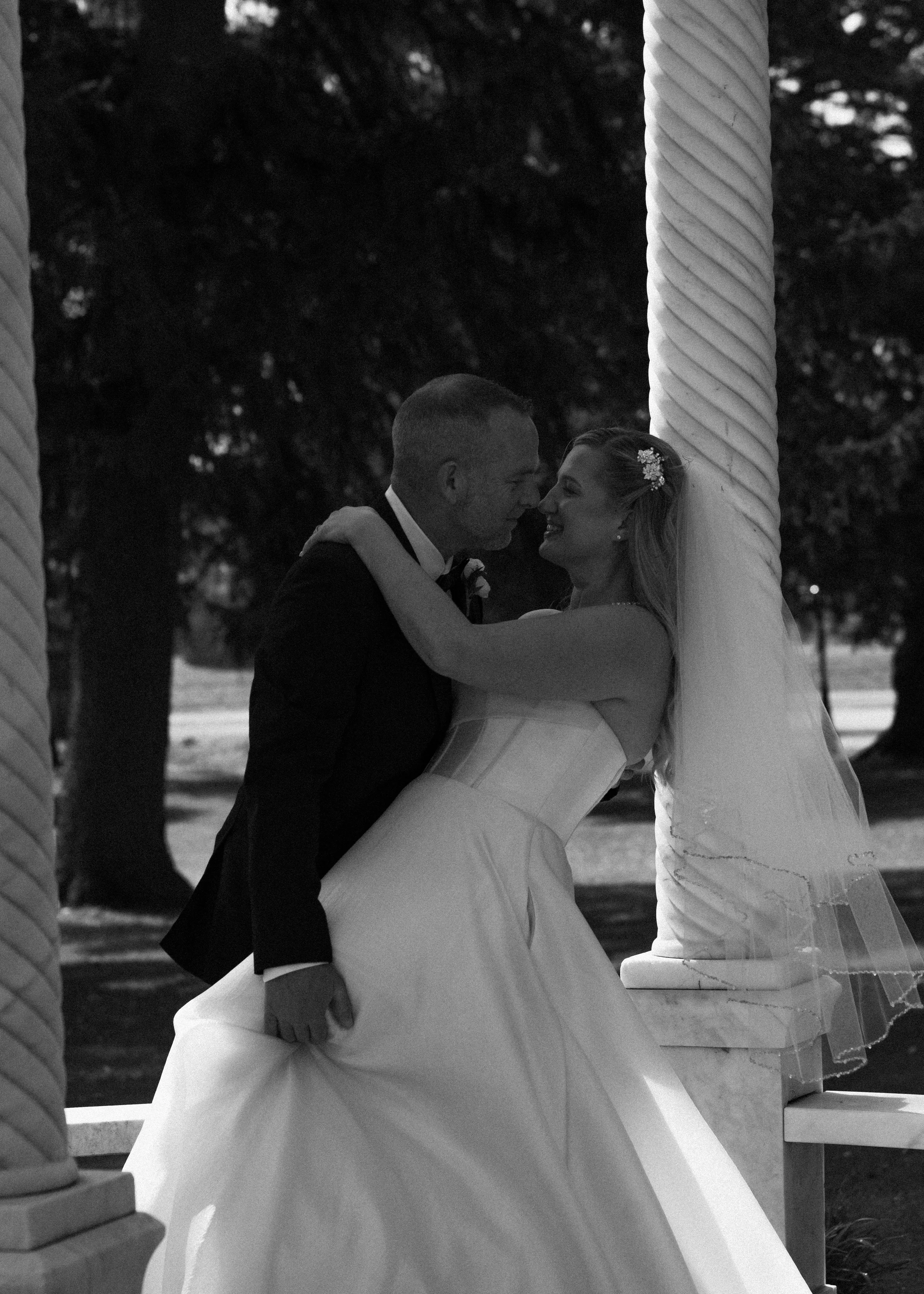 A black and white photo of a bride and groom dancing closely outdoors, with large candle holders and trees in the background.