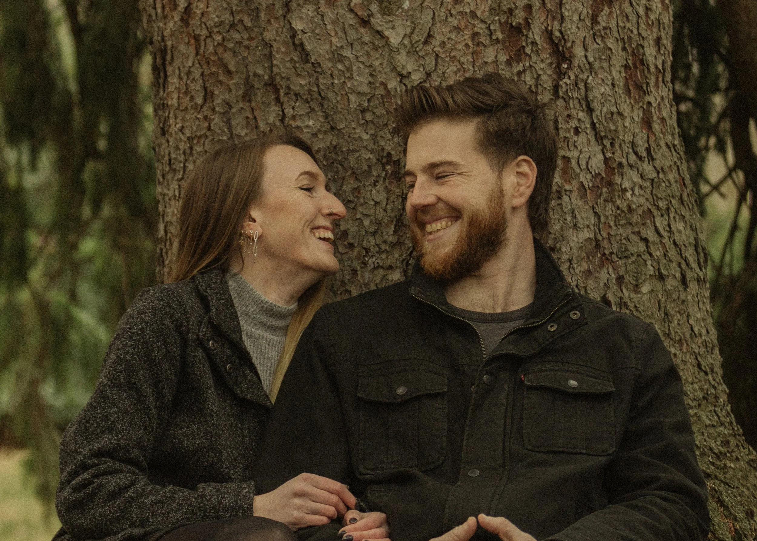A man and a woman sitting closely together outdoors in front of a tree, smiling and looking at each other.