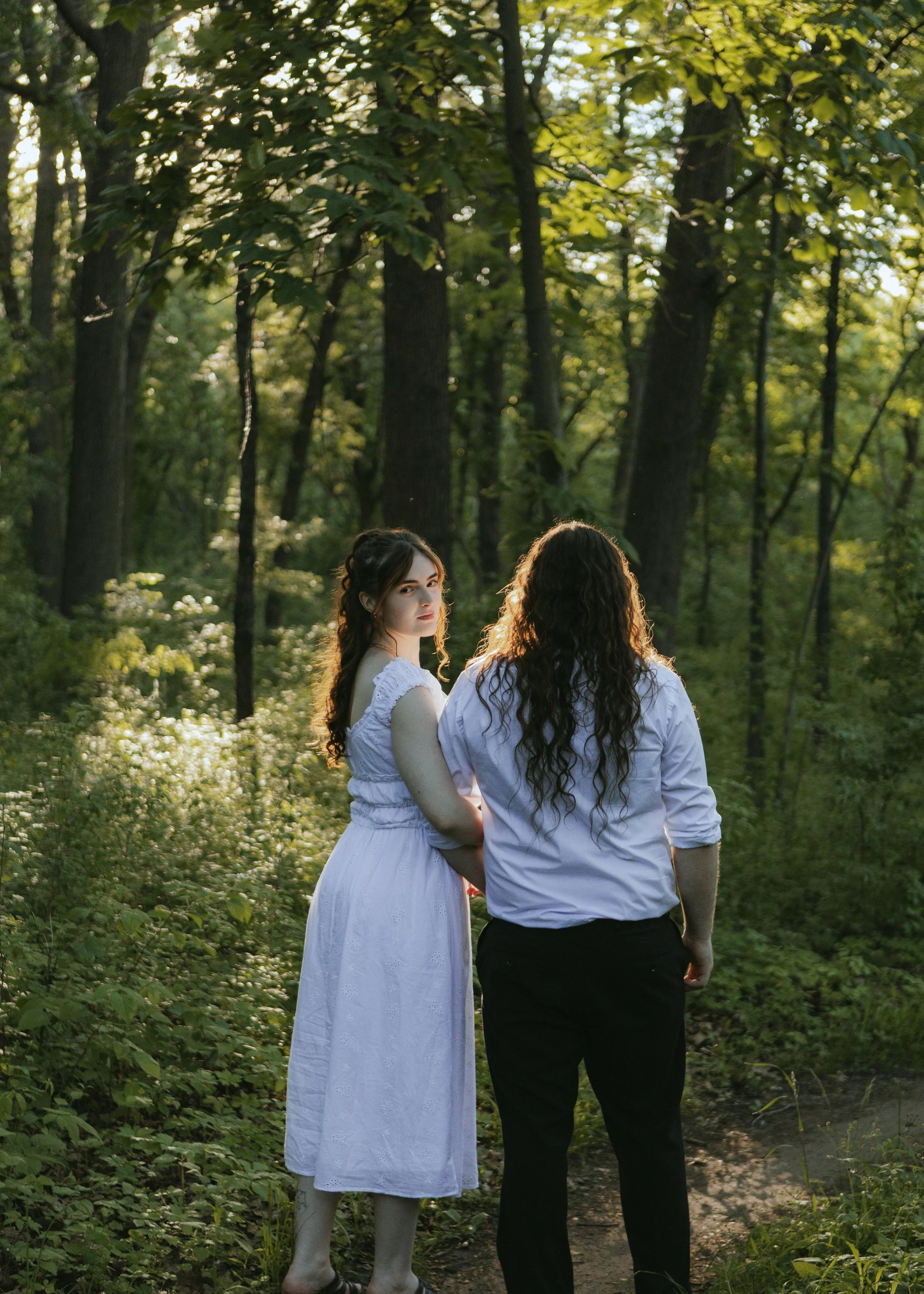 Two women standing in a lush green forest with sunlight filtering through the trees. One woman faces the camera with long, curly hair and a white dress, while the other woman, with long dark hair and a white shirt, faces away from the camera. The scene appears peaceful and natural.