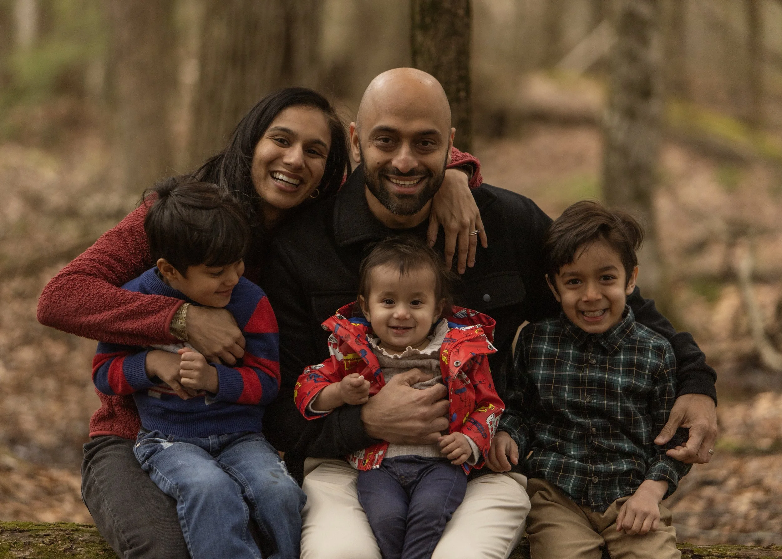 A diverse group of adults and children smiling and embracing outdoors in a wooded area during fall.
