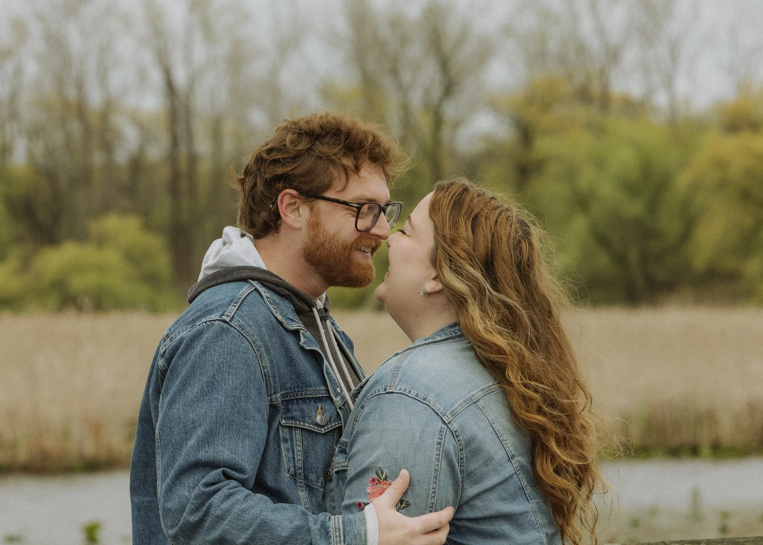 A couple smiling and looking into each other's eyes outdoors during fall, wearing denim jackets.