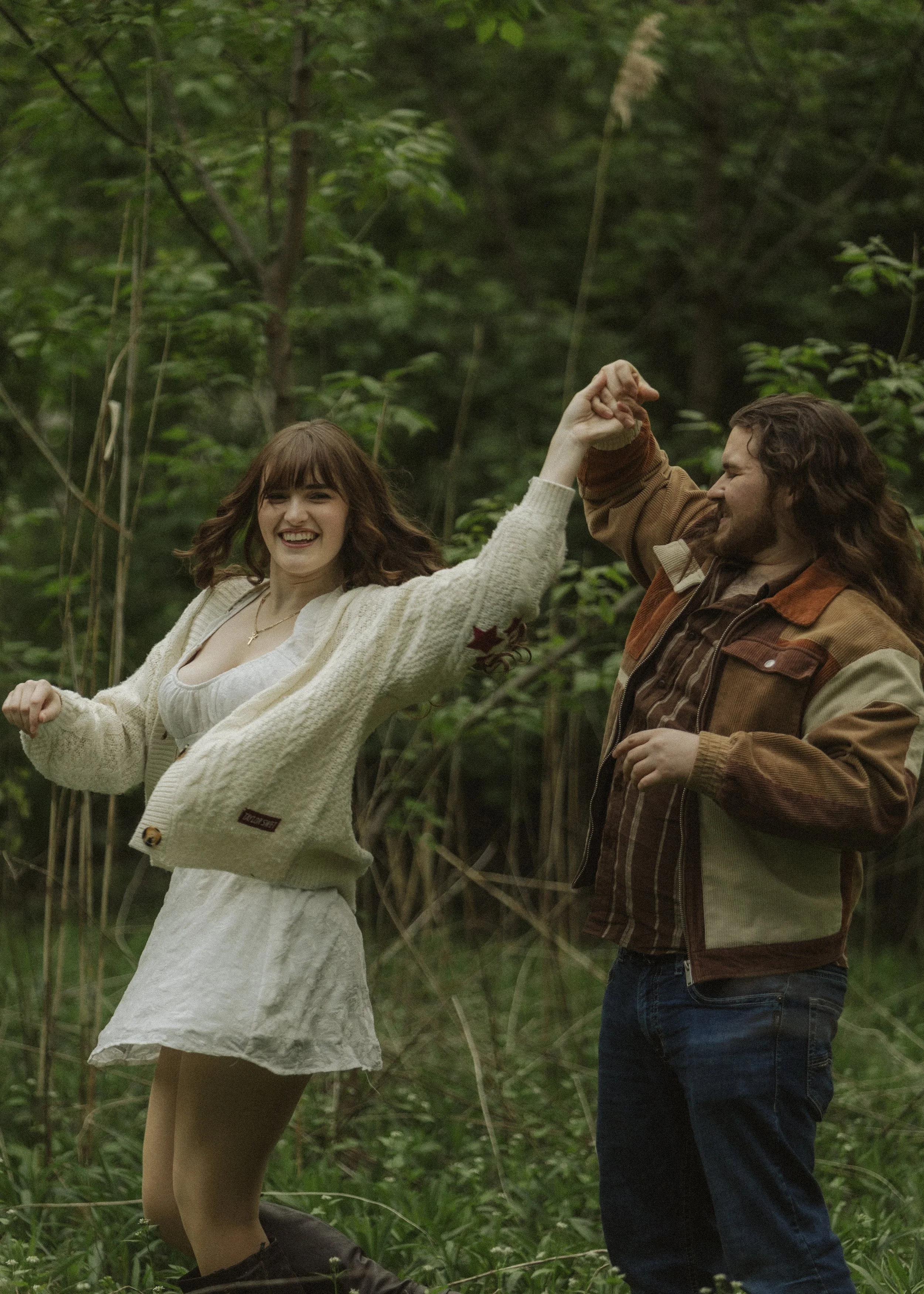A young woman in a white dress and a beige cardigan is smiling and dancing with a man wearing a brown and beige jacket. They are holding hands and appear to be enjoying themselves in a wooded area.