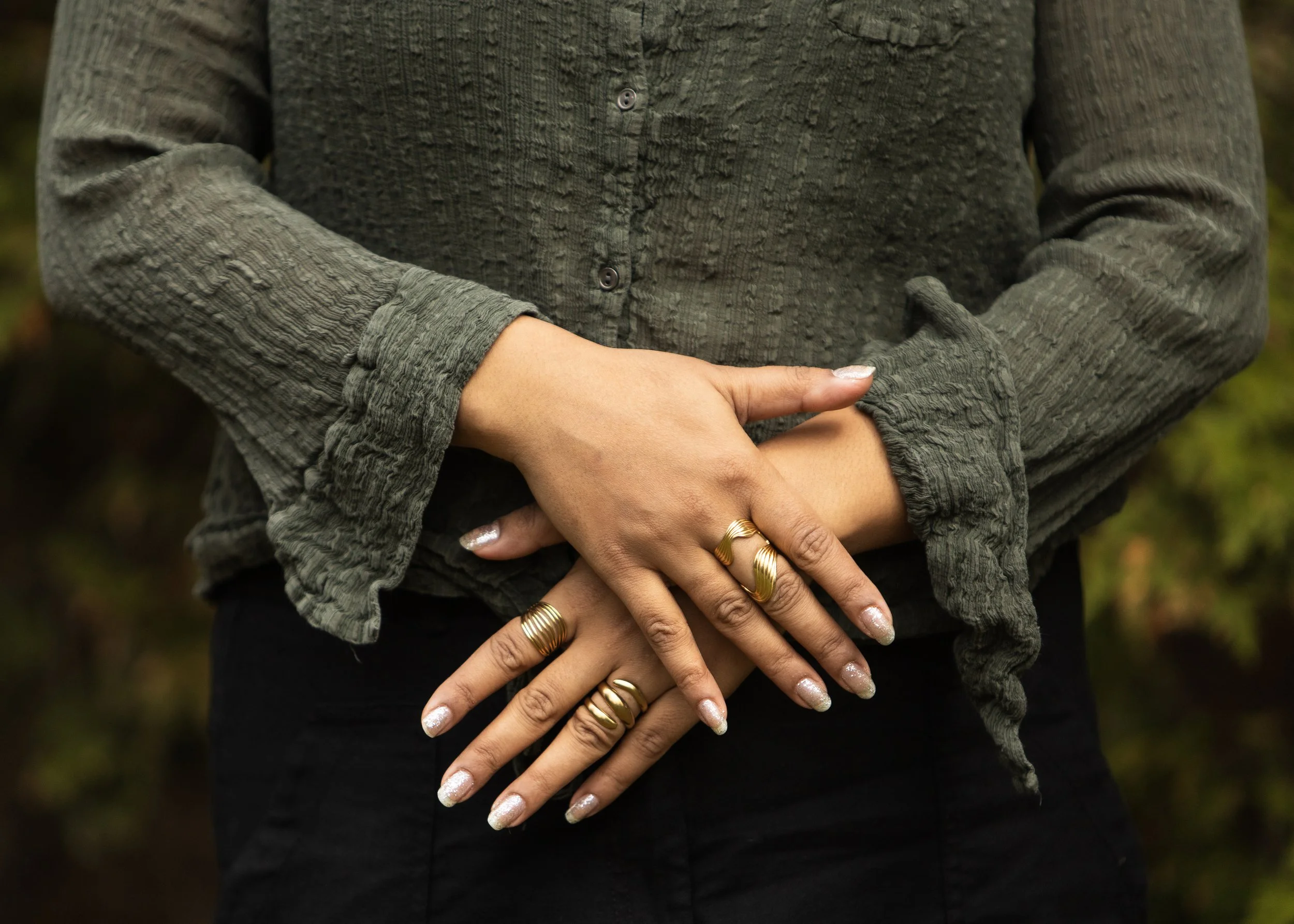 Person standing outdoors with arms crossed over their abdomen, wearing a green textured shirt and multiple gold rings on fingers.