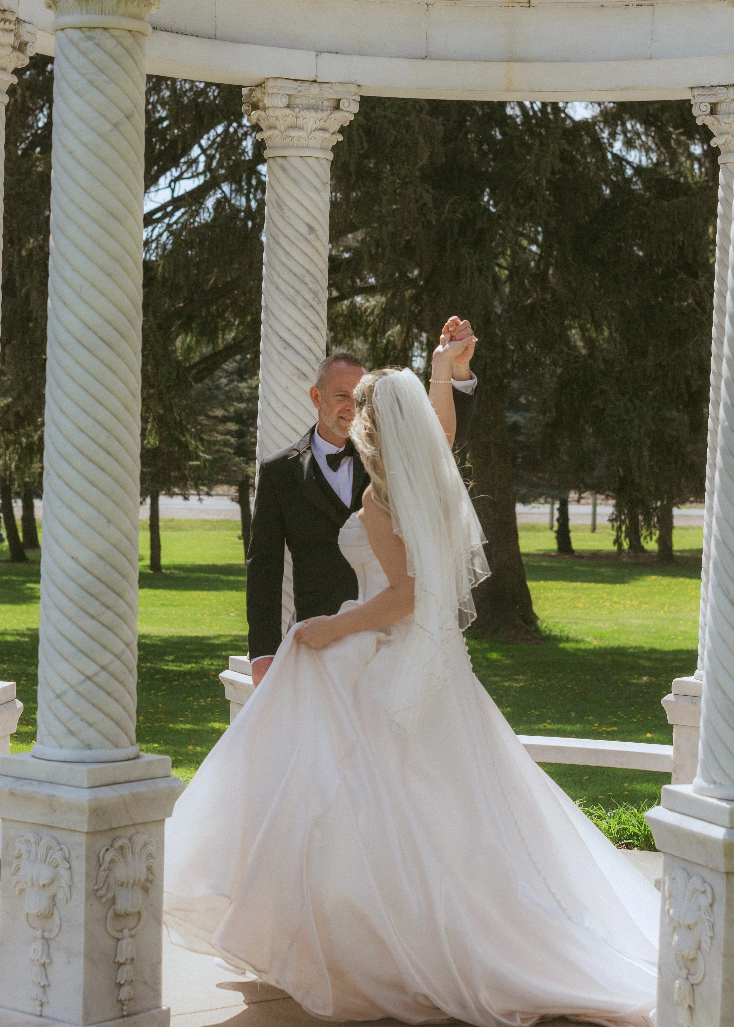 A bride and groom dancing outdoors under a white pavilion with ornate columns, green grass, and trees in the background.