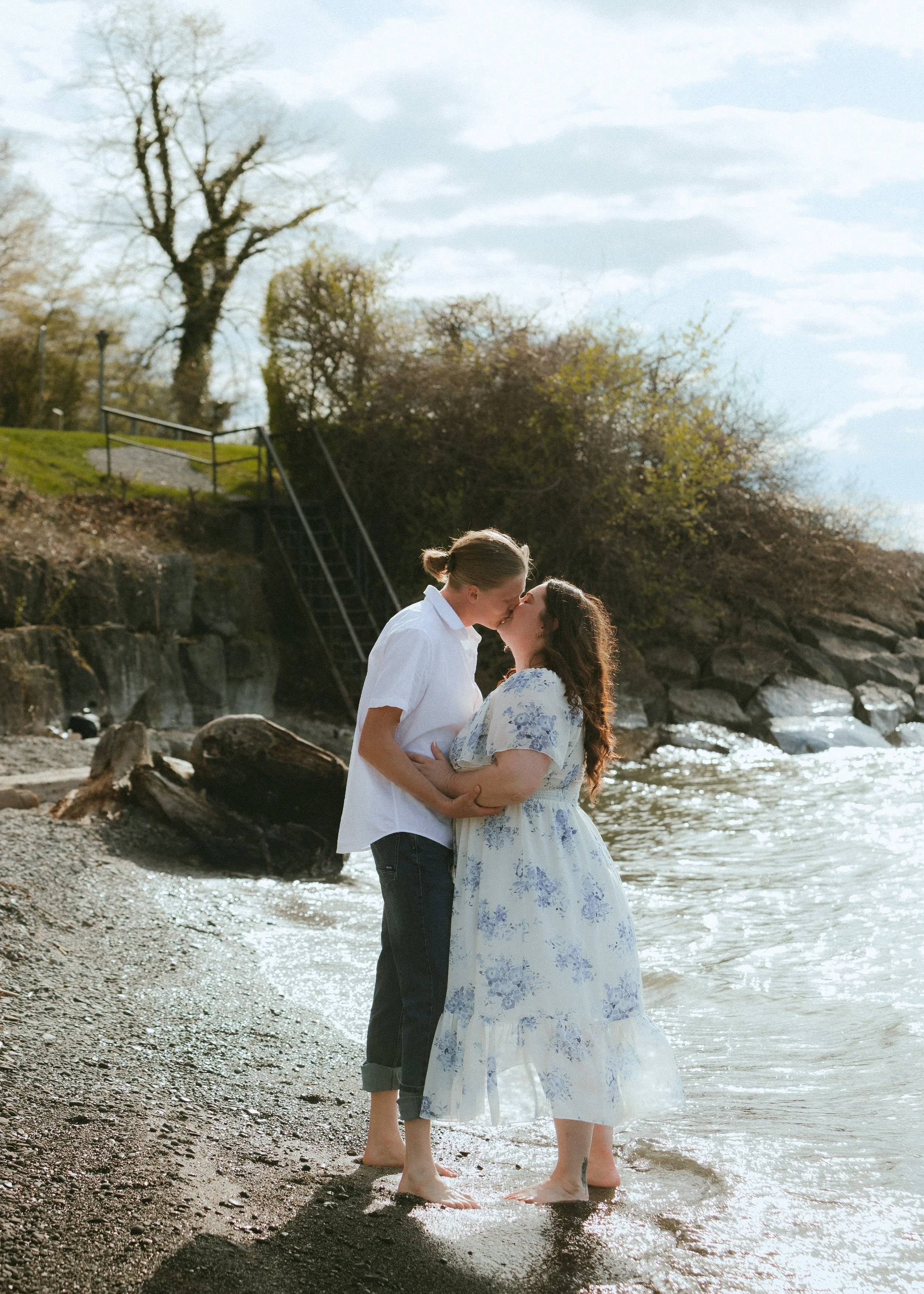 A couple embraces and kisses at the edge of a body of water, with trees and stairs in the background on a partly cloudy day.