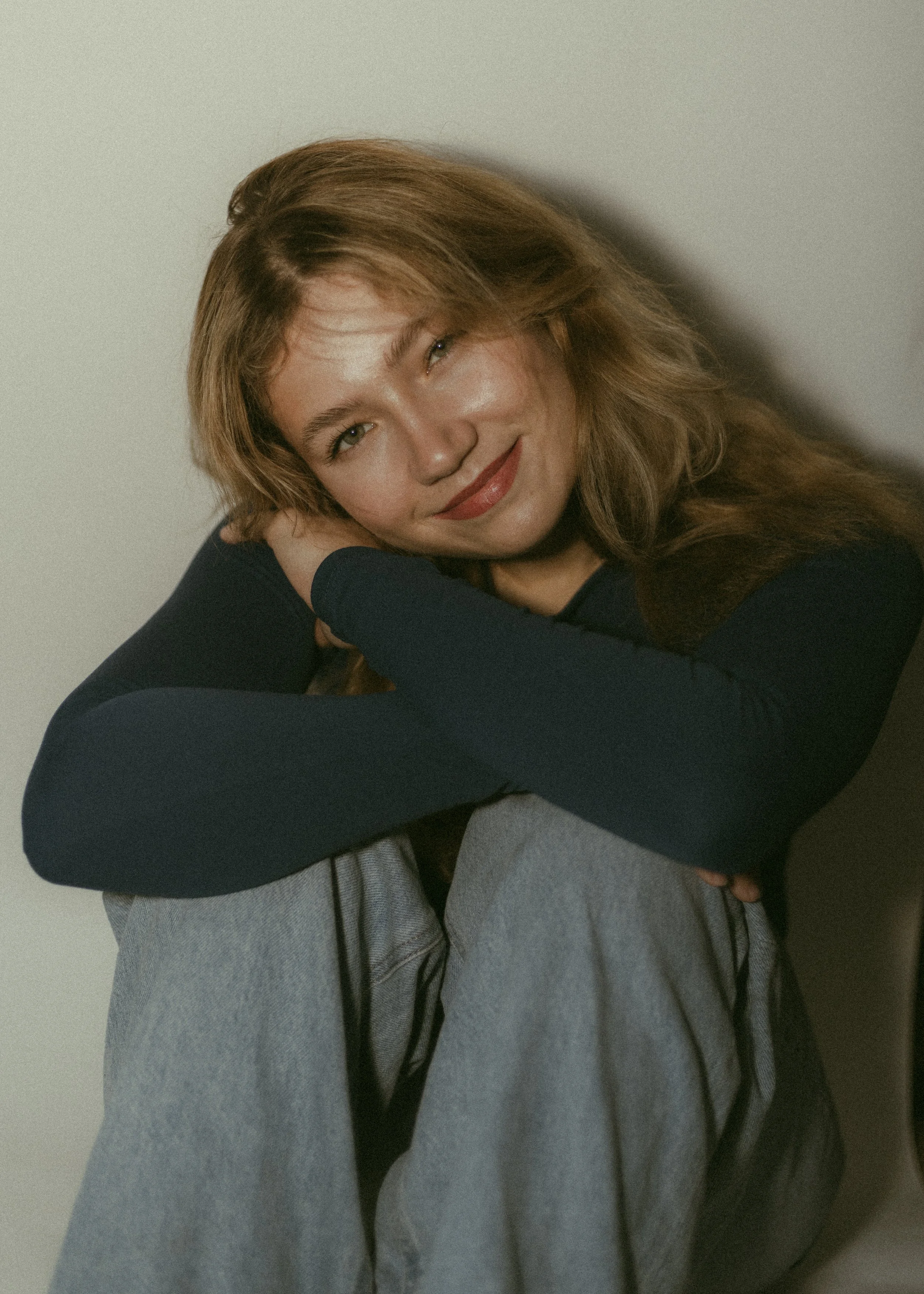 A woman with long, wavy, light brown hair smiling and leaning her head on her hand. She is wearing a black long-sleeve top and gray pants, sitting against a plain, light-colored background.