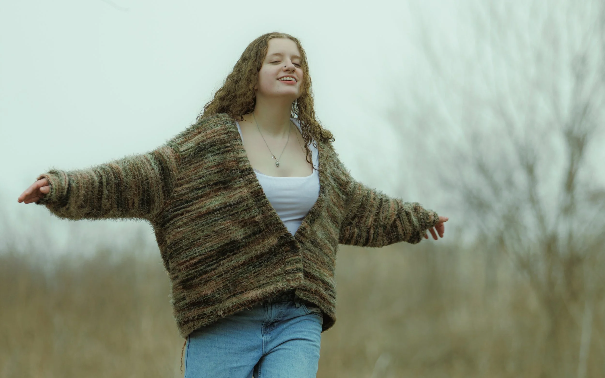 A young woman with long curly hair enjoying outdoors in a field with her arms outstretched