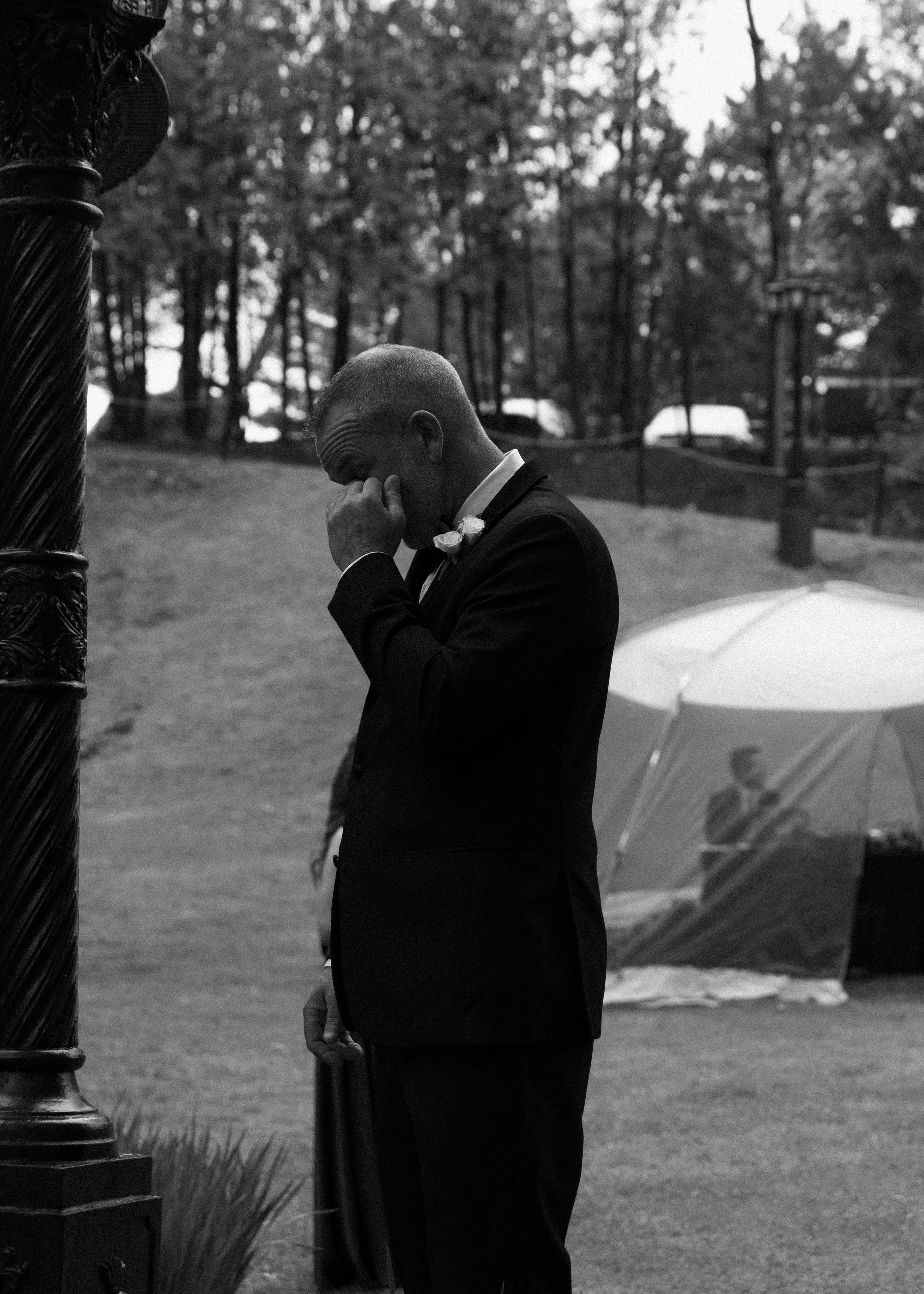 A man dressed in a suit with a boutonniere is standing outdoors, wiping his face with his hand. He appears to be emotional or distressed. There is an umbrella in the background, and a decorative post is partially visible on the left.