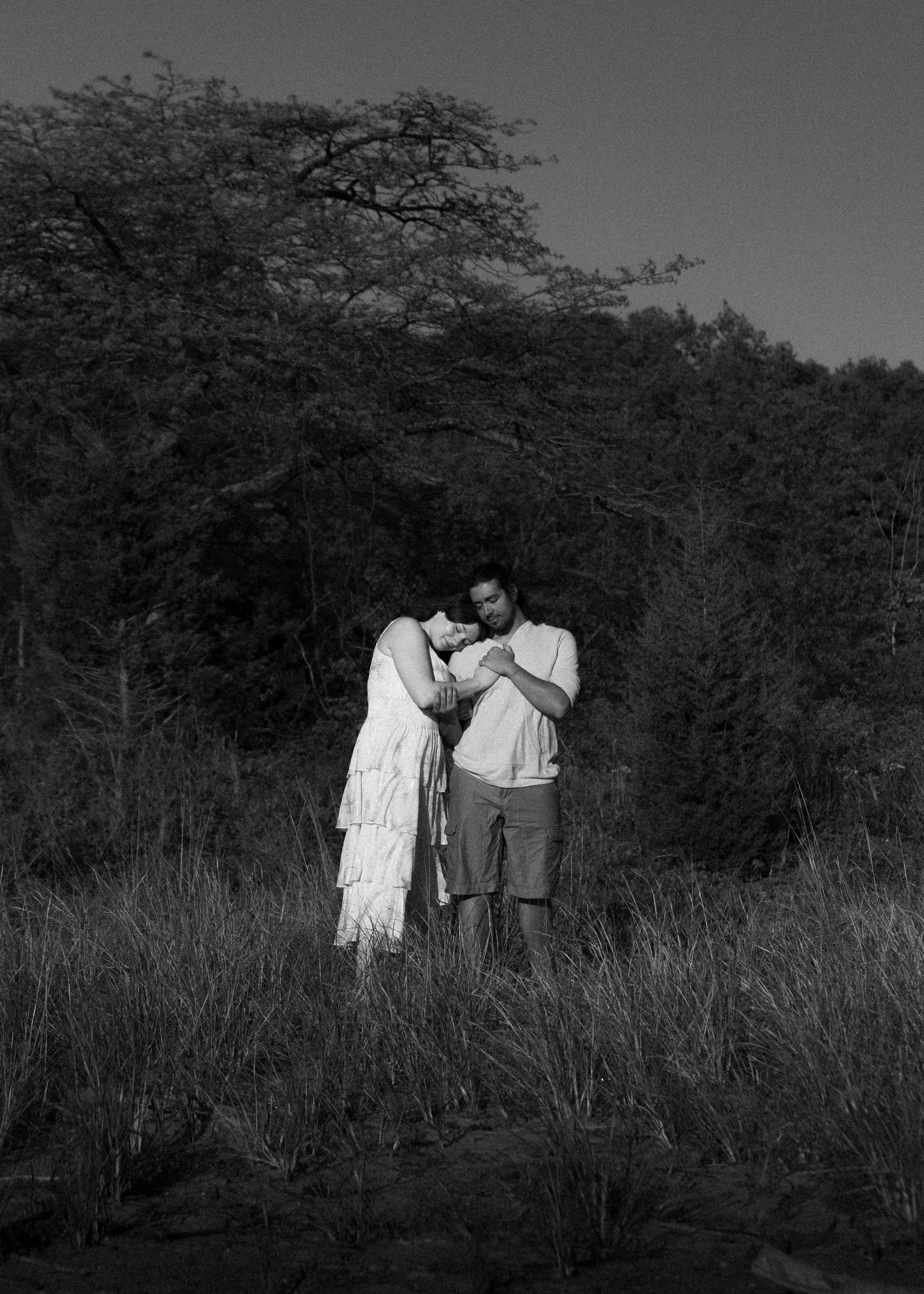 A black and white photo of a couple standing in a field with trees in the background. The woman has short hair and is wearing a sleeveless dress, leaning her head on the man's shoulder. The man has a beard and is wearing a long-sleeved shirt rolled up to the elbows and shorts. They are holding hands and looking down, with a peaceful, intimate atmosphere.