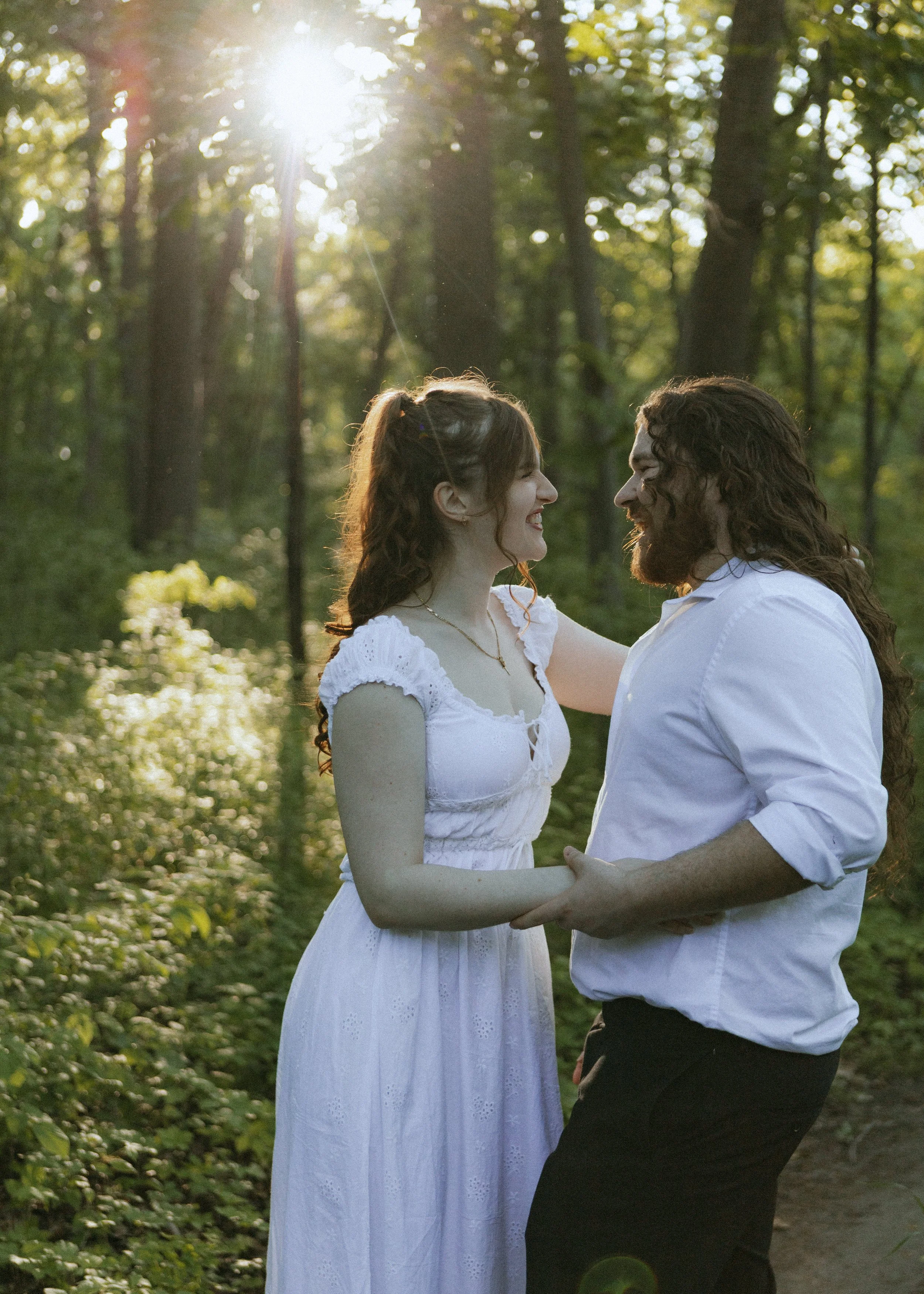 A couple is standing close together outdoors, holding hands, and smiling at each other in a forest with sunlight filtering through the trees.