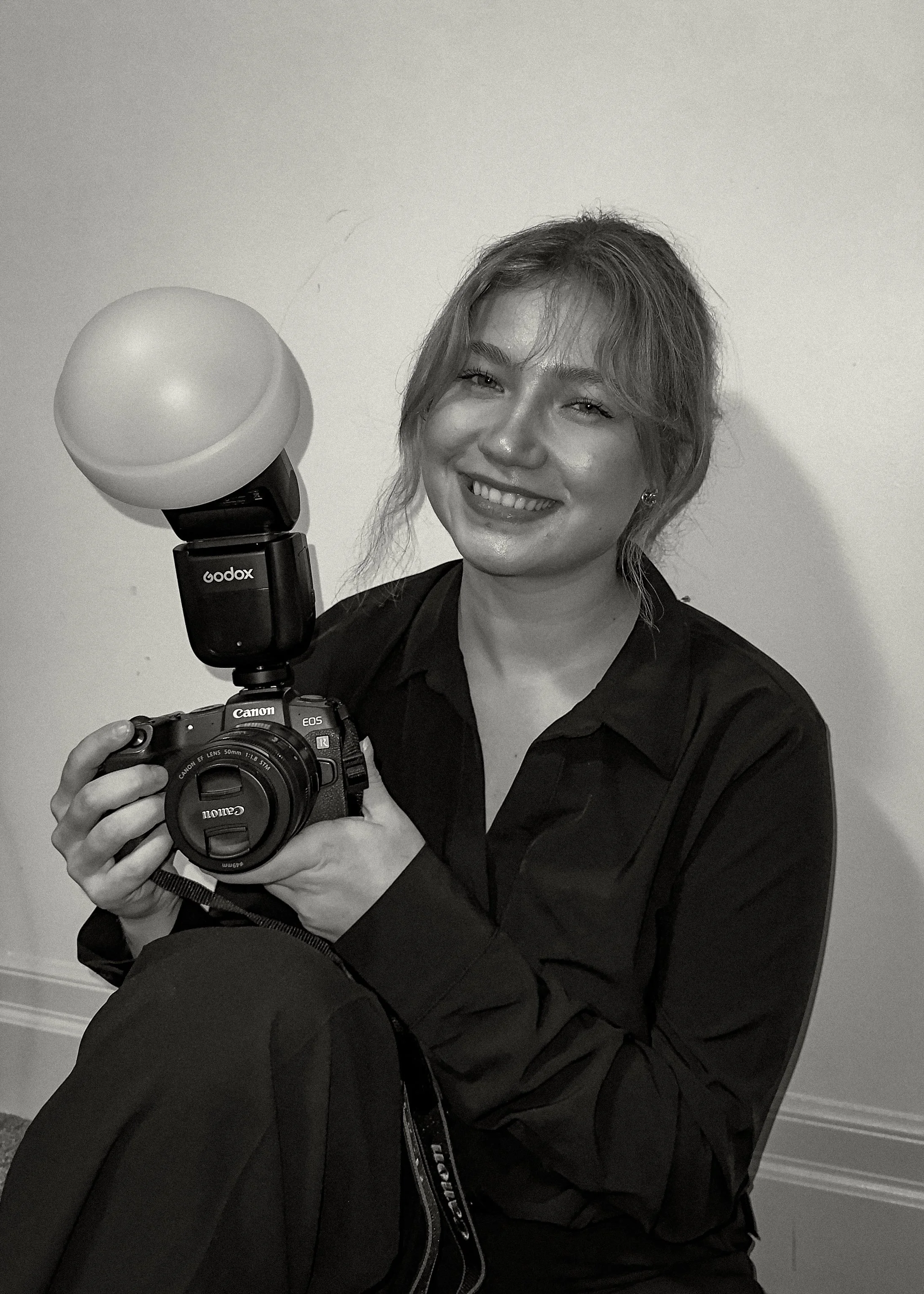 A woman with wavy hair smiling, holding a Canon camera with a large softbox light attached, sitting against a plain wall.