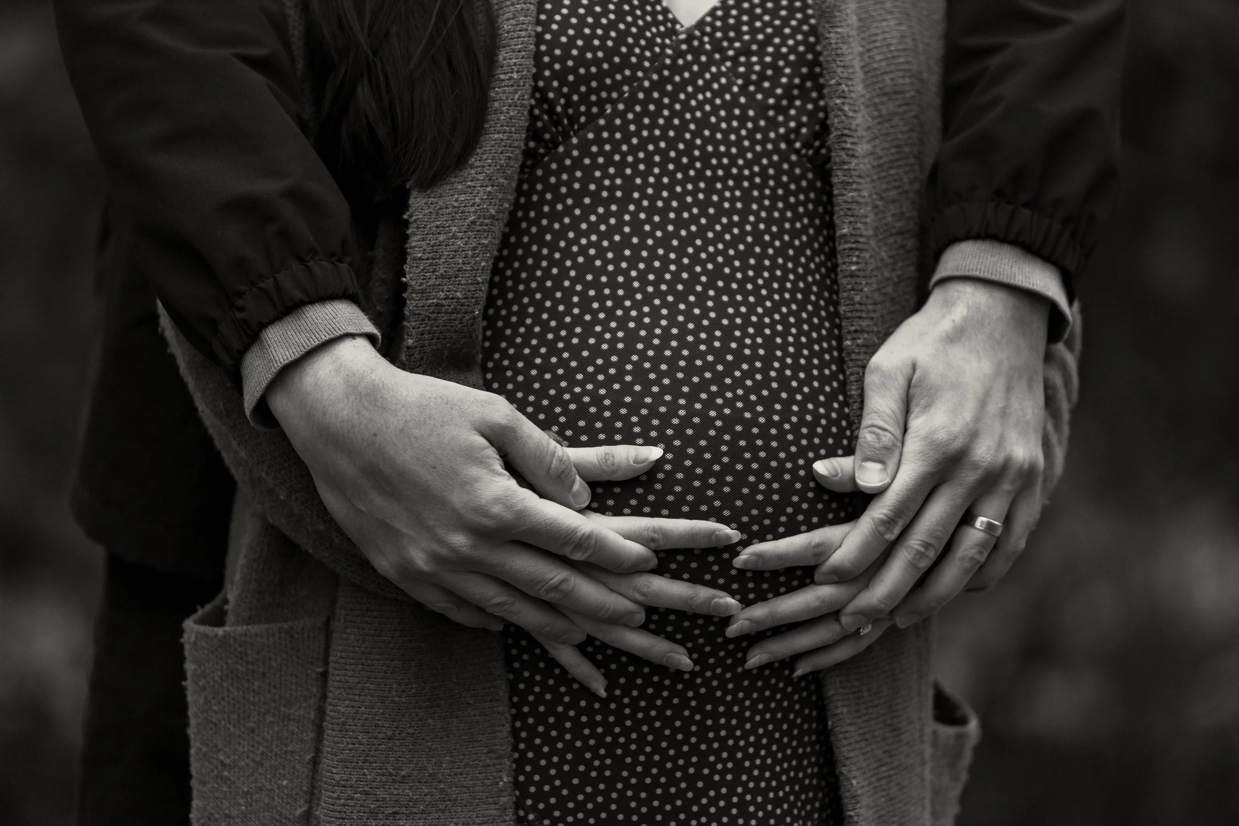 A person with dark hair wearing a black jacket and a gray knitted cardigan, holding their pregnant belly with both hands. The person is dressed in a black and white polka dot top.