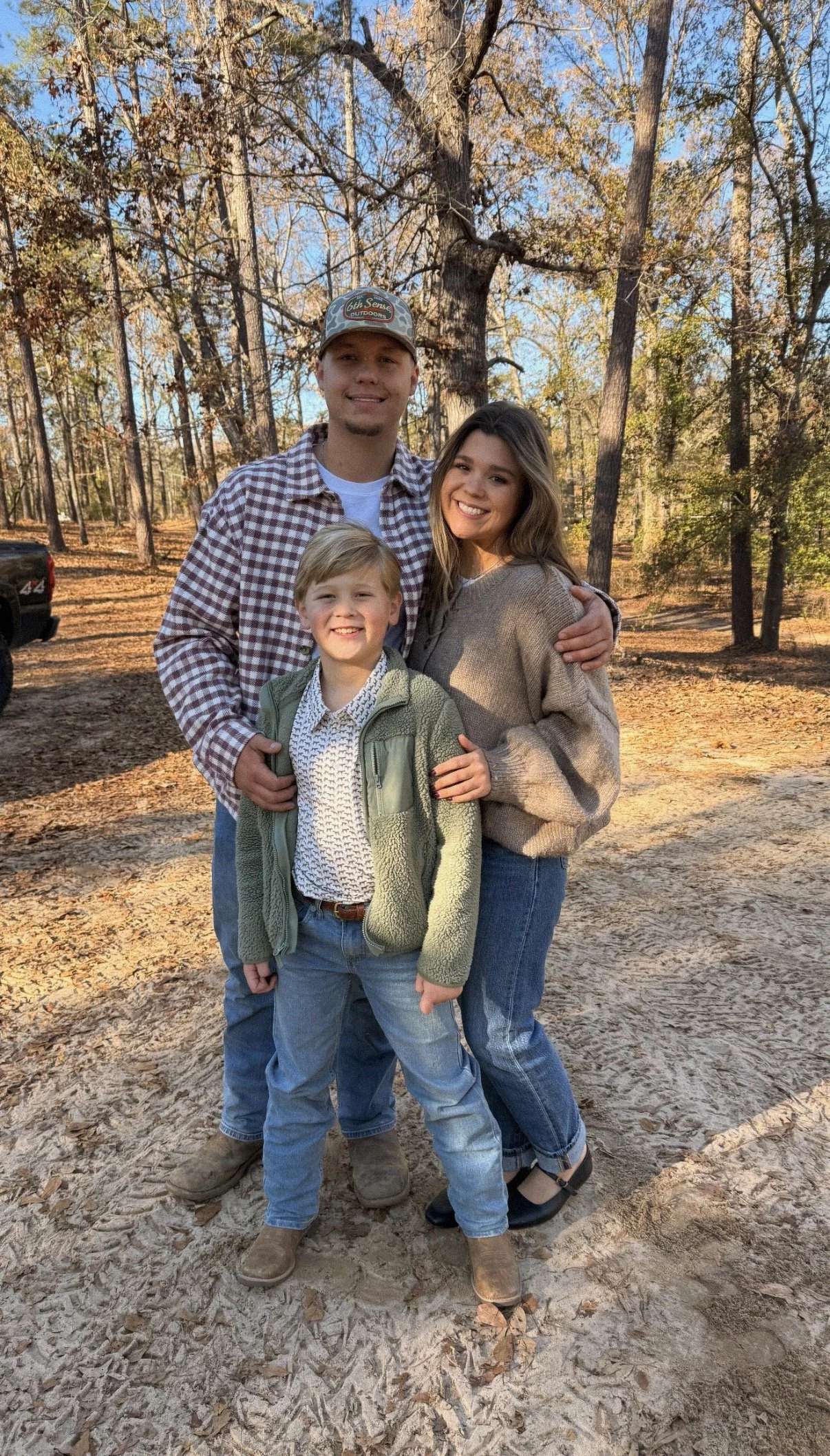 A family of three posing outdoors in a forested area during autumn, with trees and fallen leaves in the background.
