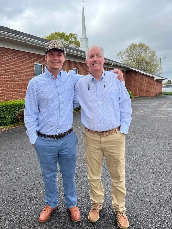 Two men standing outdoors in front of a brick church with a steeple, smiling and posing together. One is younger, wearing a light blue dress shirt, blue pants, and brown shoes; the other older, wearing a light blue dress shirt, beige pants, and tan shoes.