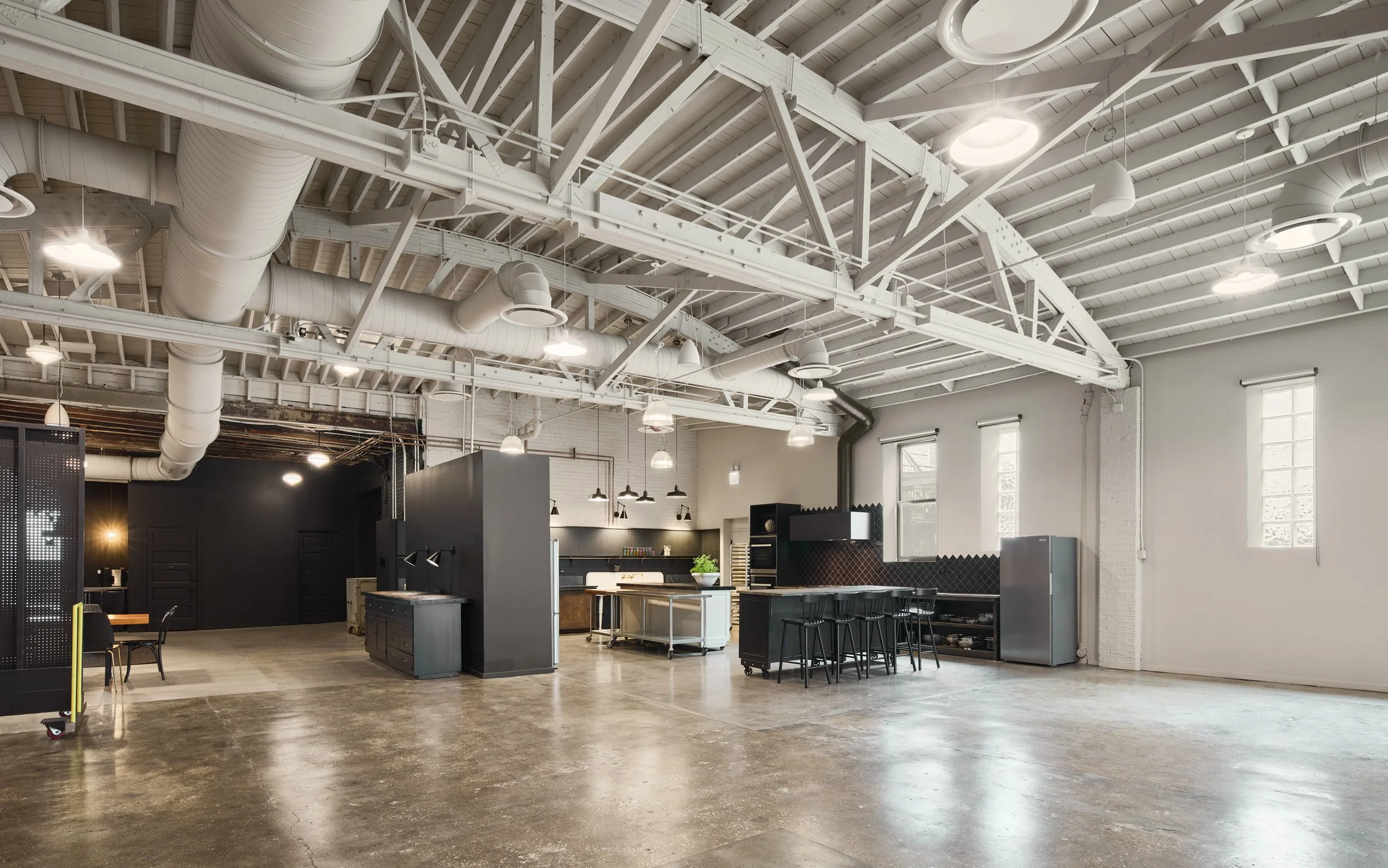 Open plan office with white exposed ceiling beams, ductwork, large windows, and black and white furniture.