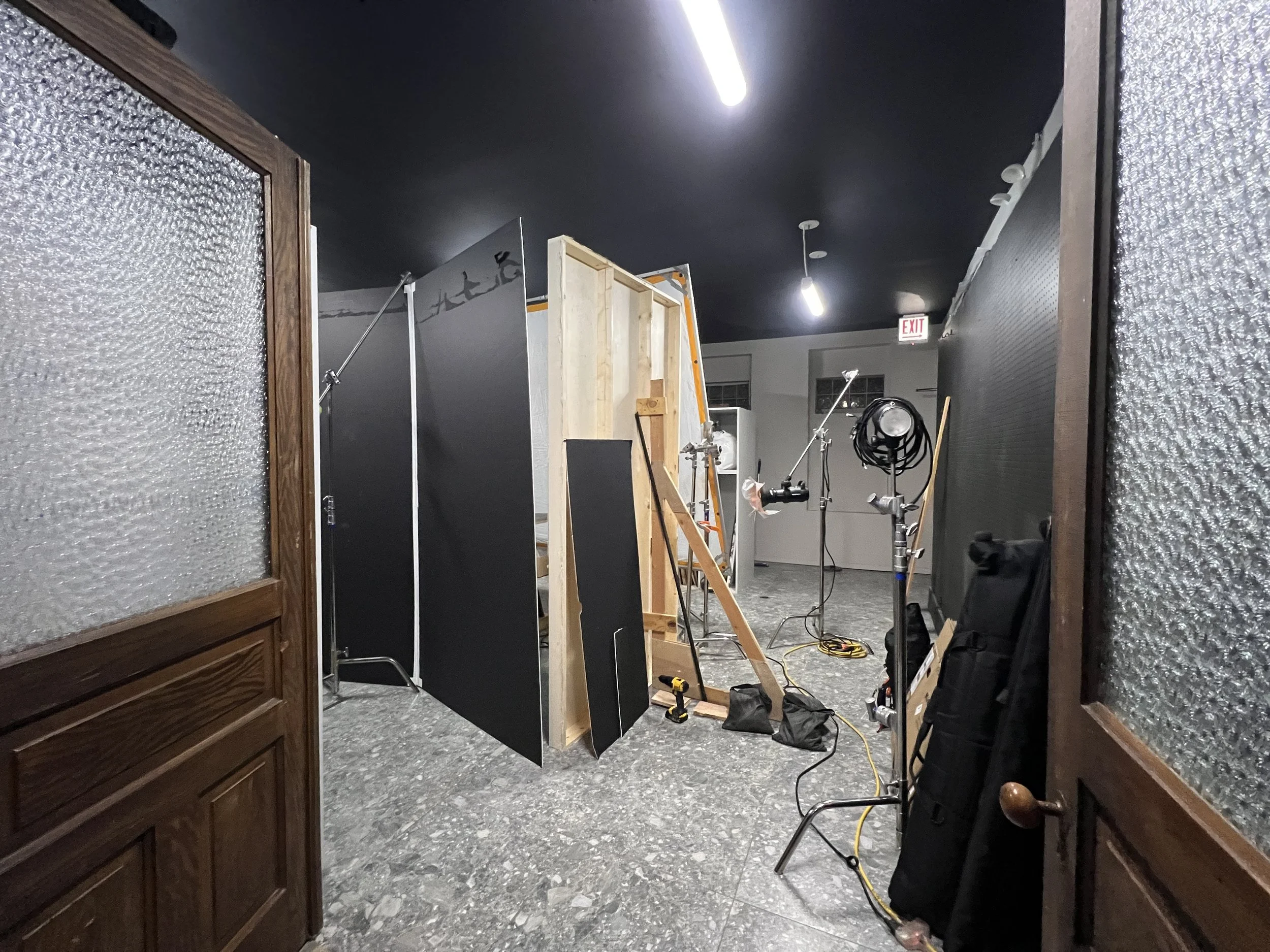 Interior of a photography or film studio with lighting equipment, a black backdrop, a ladder, and construction materials. The space is viewed through an open door with frosted glass panels.