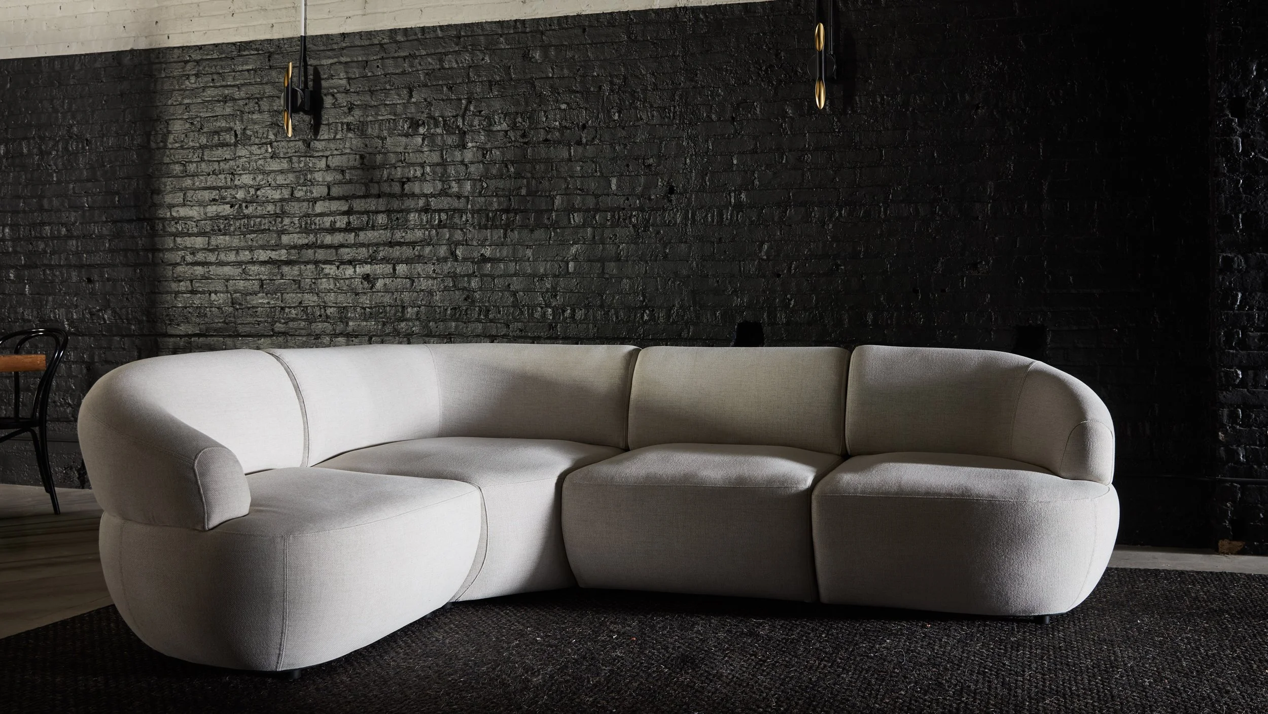A cream-colored, L-shaped sofa with rounded edges in front of a black brick wall, accompanied by a small wooden side table and a black chair with a wooden armrest.