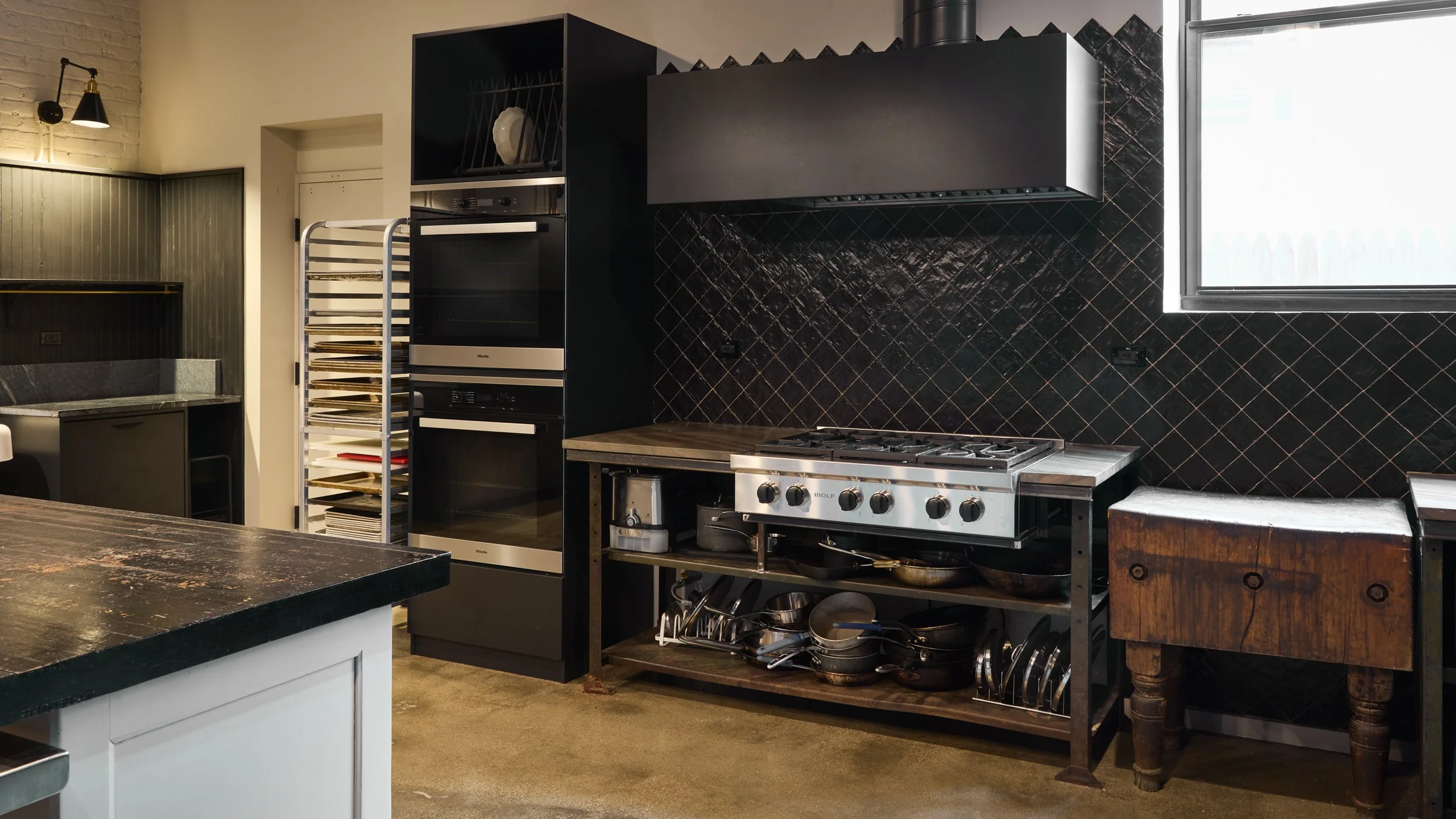 A modern kitchen with black and dark gray cabinetry, a black tile backsplash, a stainless steel stove, and a window. There is a vintage wooden table to the right, and various kitchen utensils on a lower shelf.
