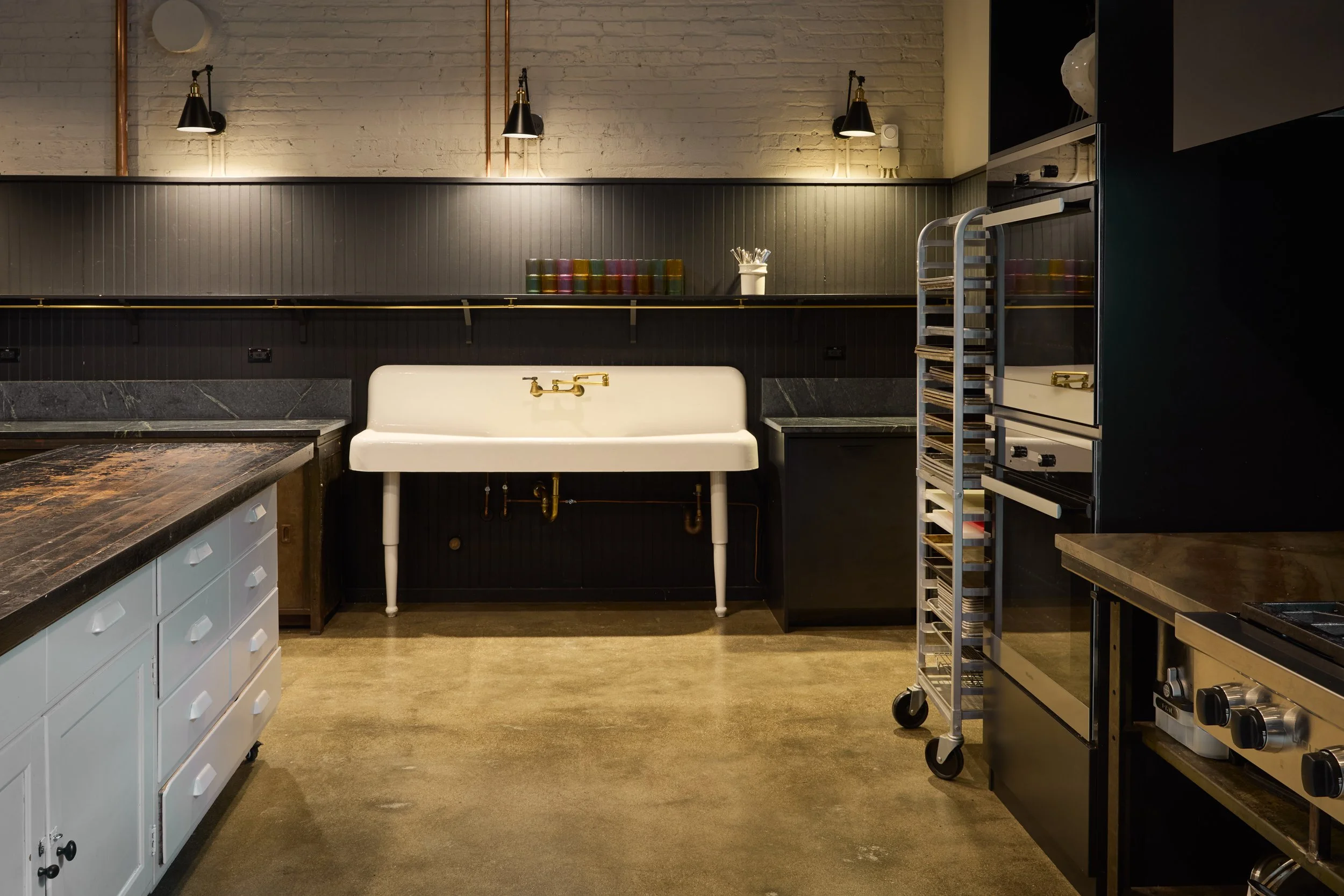 An industrial-style kitchen with black and white cabinetry, a white sink with brass fixtures, a black wall with vertical paneling, and metal shelving units on wheels.