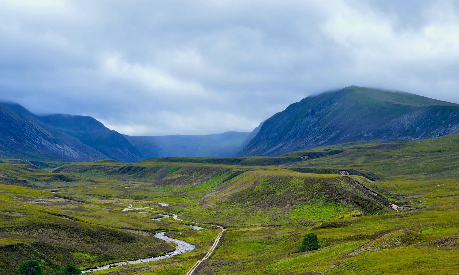 Scenic landscape of green rolling hills with a river flowing through, surrounded by mountains under cloudy sky.