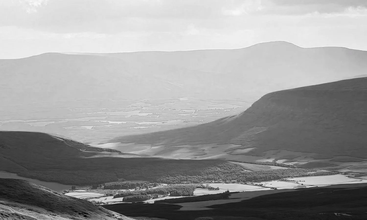 A black and white landscape photo of rolling hills, fields, and valleys with mountains in the background under an overcast sky.