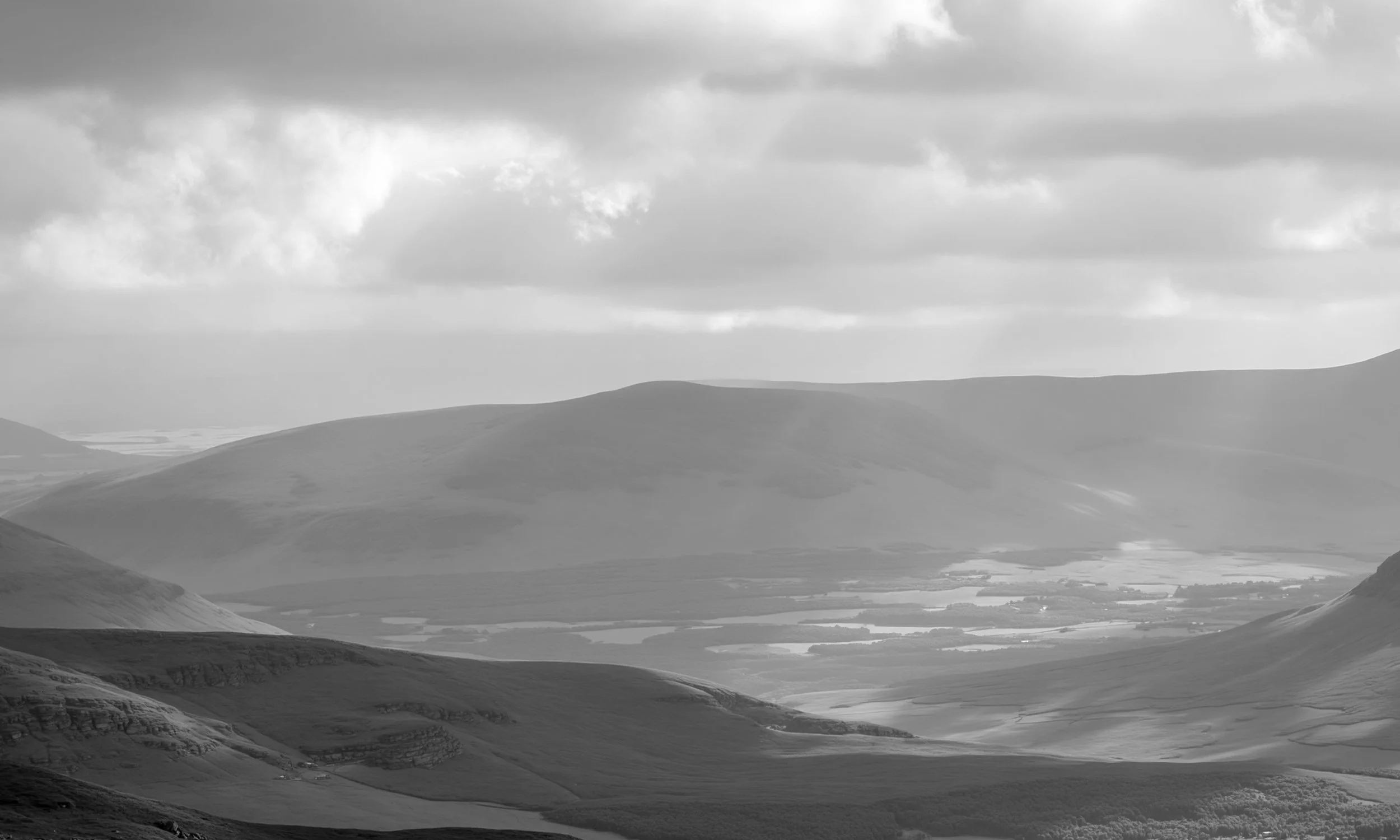 Black and white photo of rolling hills and mountains with a cloudy sky overhead, distant lakes or rivers in the valley.