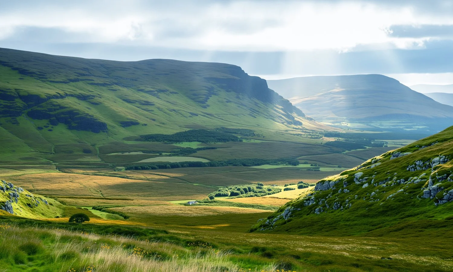 Scenic landscape of rolling green hills and mountains with patches of sunlight breaking through clouds and illuminating the valley below.