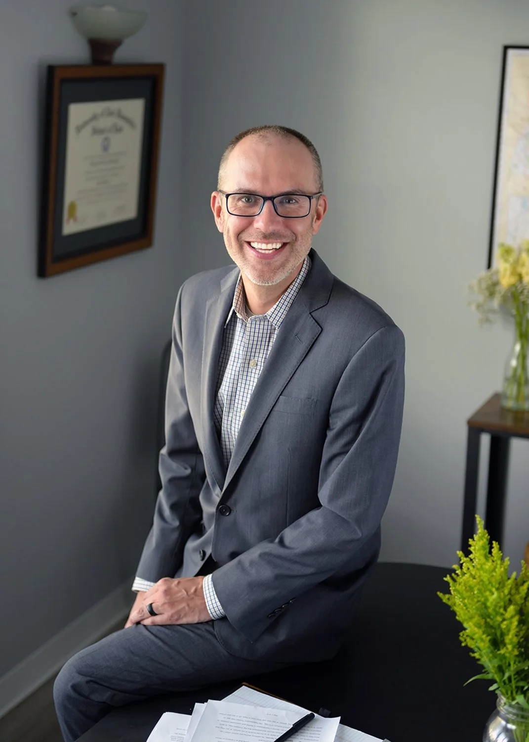 A smiling man with glasses in a gray suit is sitting on a desk in an office setting, with framed certificates on the wall and a small table with a flower arrangement in the background.