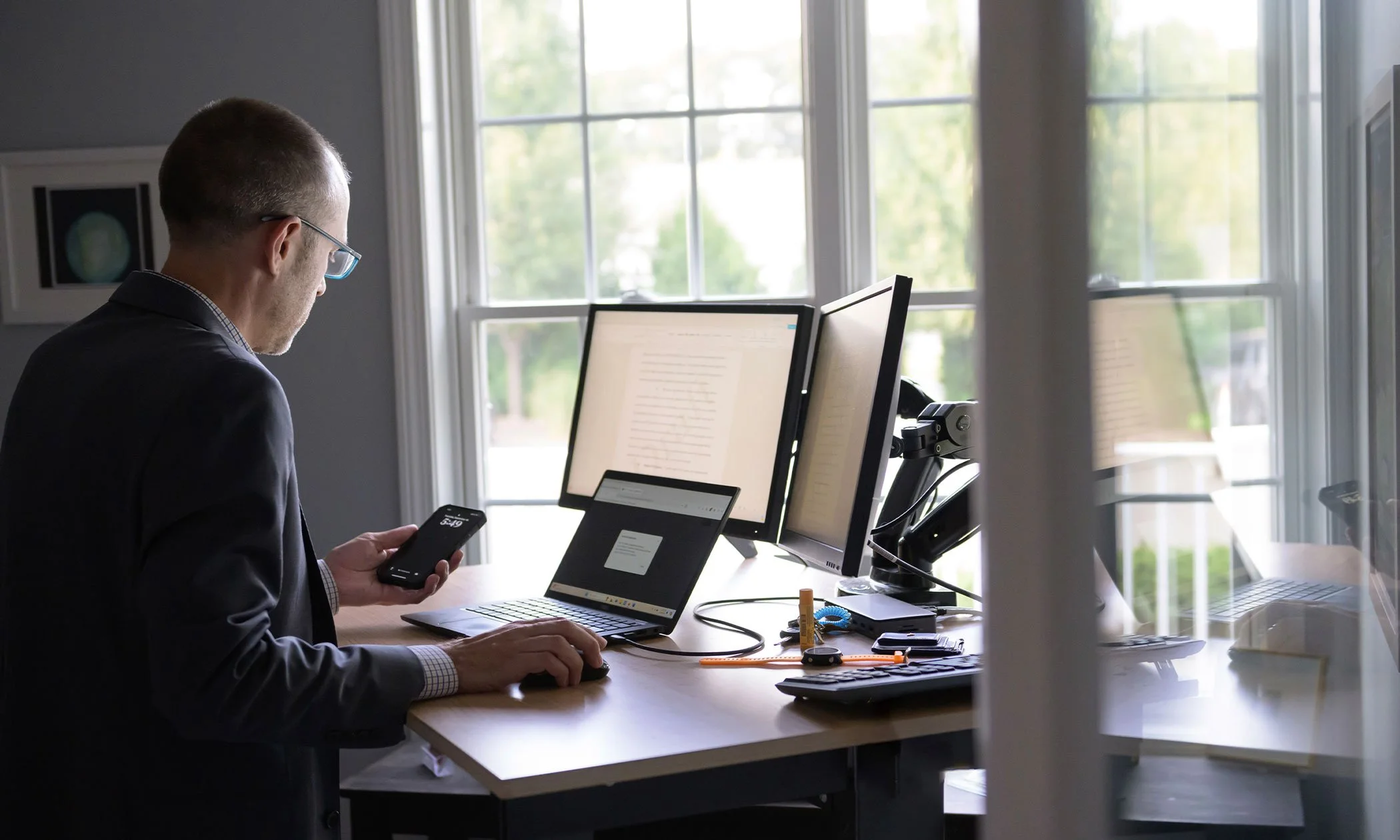 A man in a suit wearing glasses using a computer mouse and looking at his smartphone in a home office with two computer monitors, a laptop, and various office supplies on the desk, with large windows in the background.