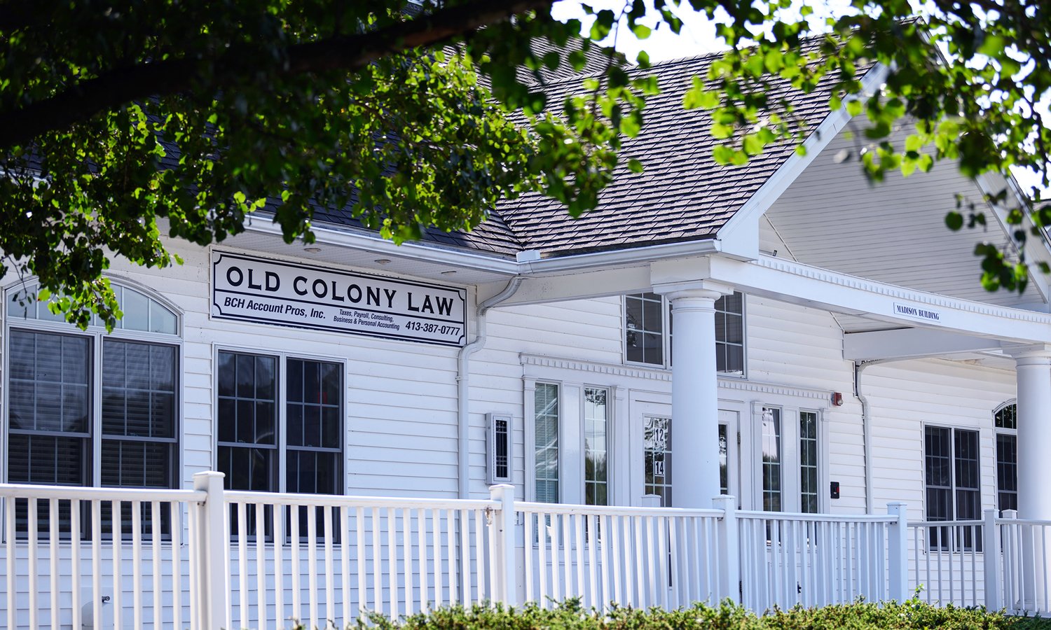 White building with a sign that reads "Old Colony Law" and features columns at the entrance, surrounded by trees and a white picket fence.