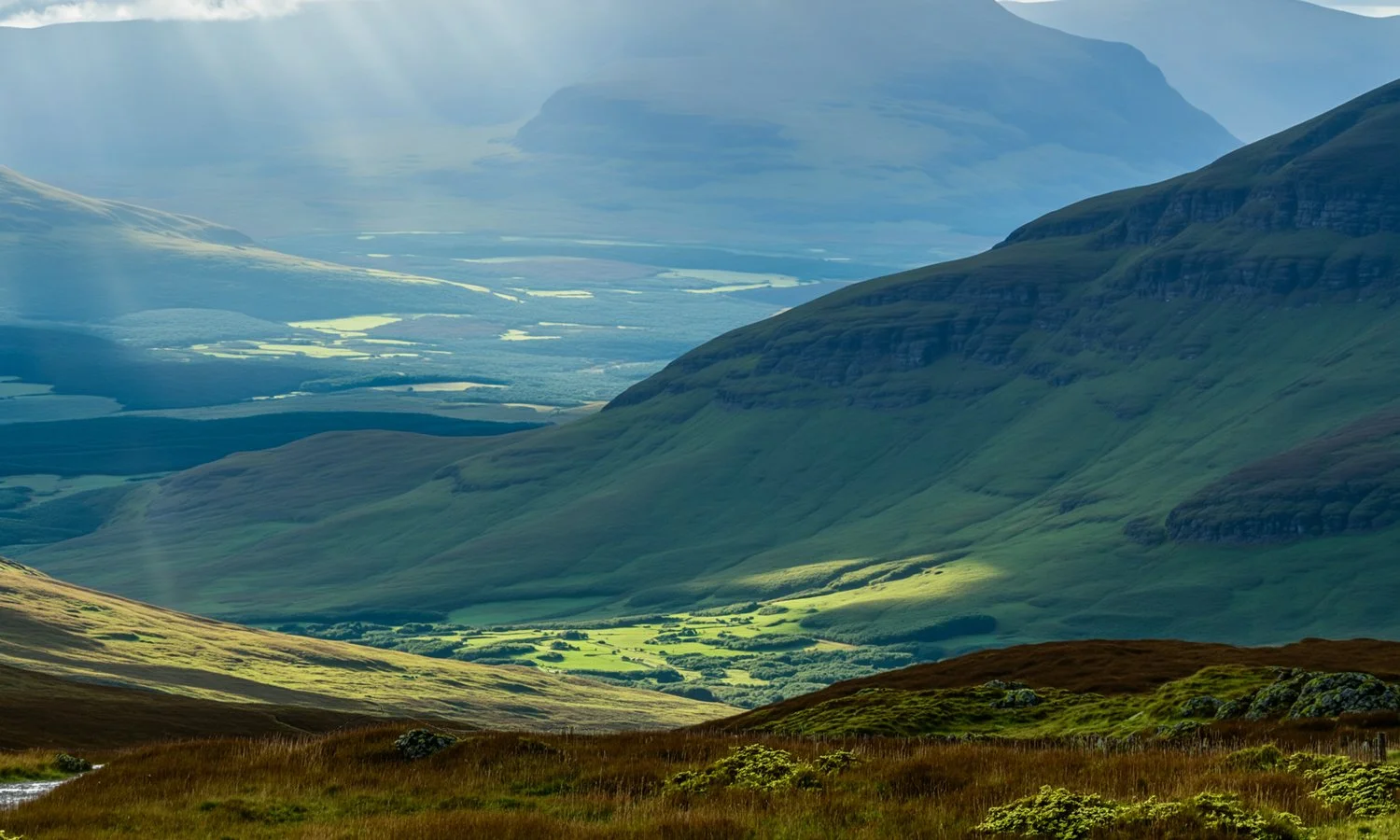 A scenic view of green rolling hills and mountains with patches of sunlight and shadow, and a river valley in the distance.