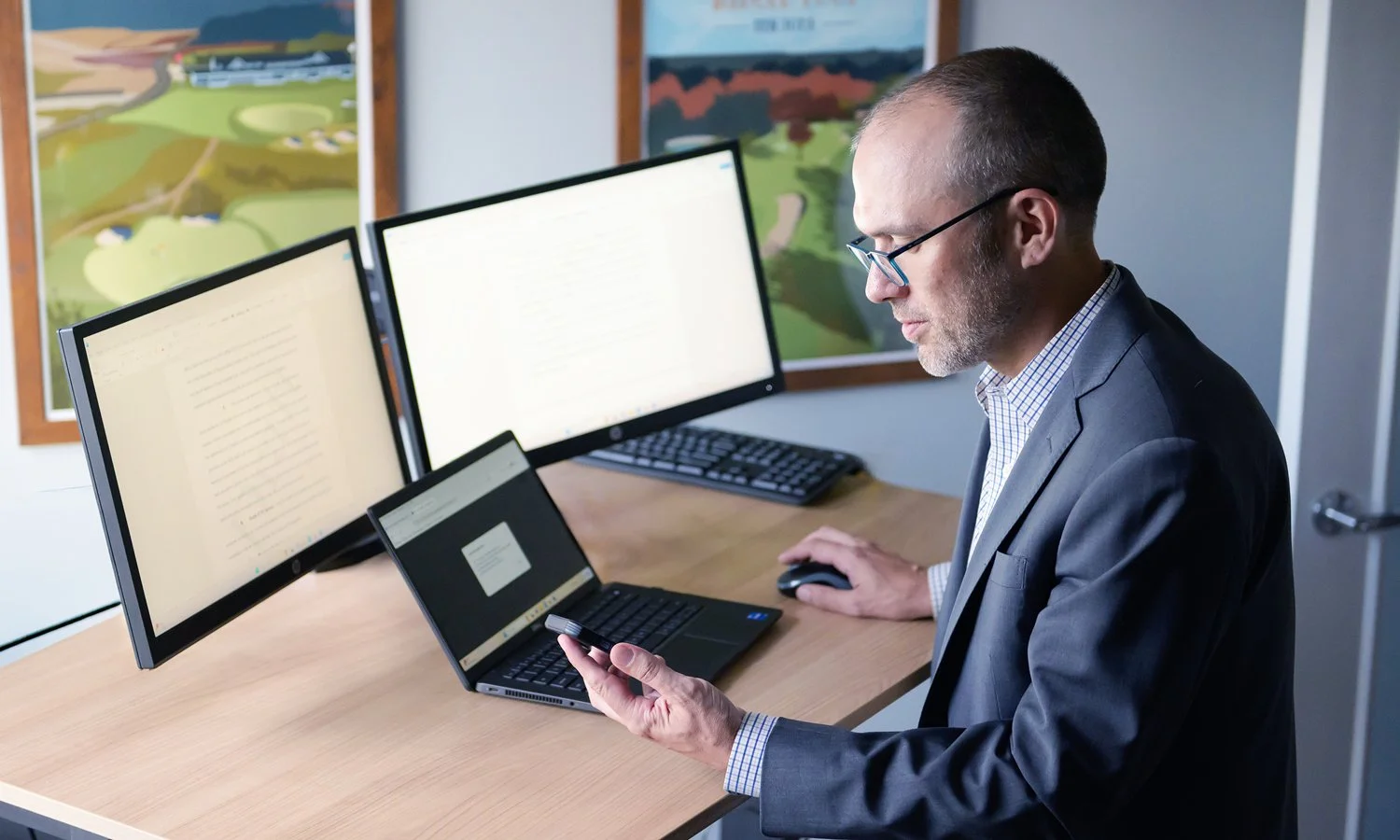 A man in a suit working at a desk with three computer monitors, a keyboard, a mouse, and a laptop, looking at his phone, with landscapes on posters behind him.