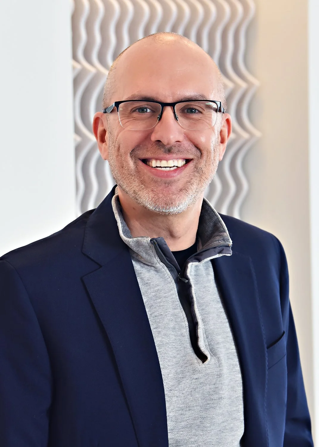 A smiling man with glasses, a gray beard, and a bald head, wearing a navy blazer over a gray zip-up sweater, standing indoors in front of a textured wall.