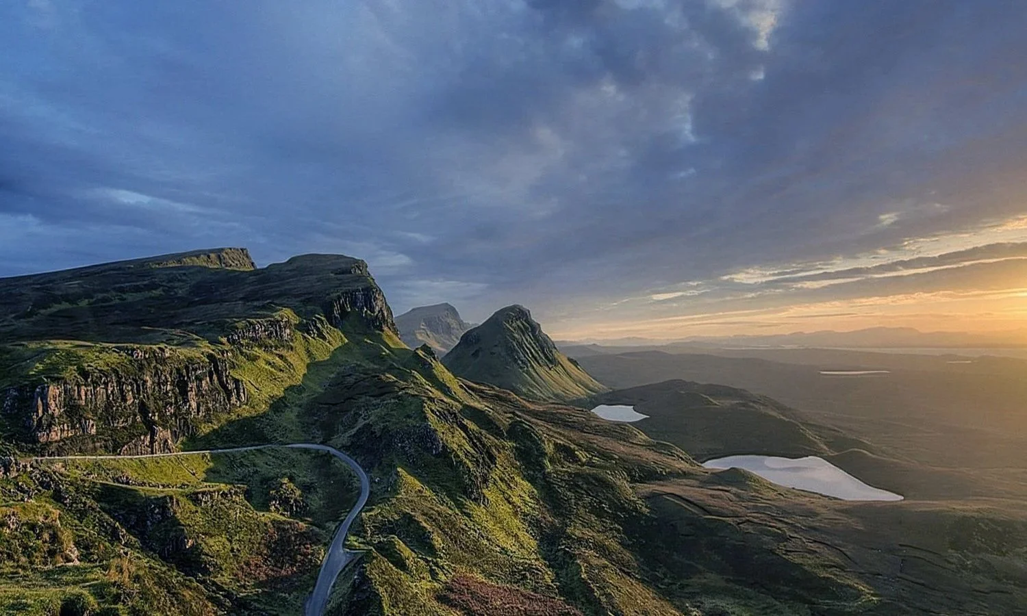 Scenic view of green, rocky mountains with a winding road, lakes, and a partly cloudy sky during sunset or sunrise.