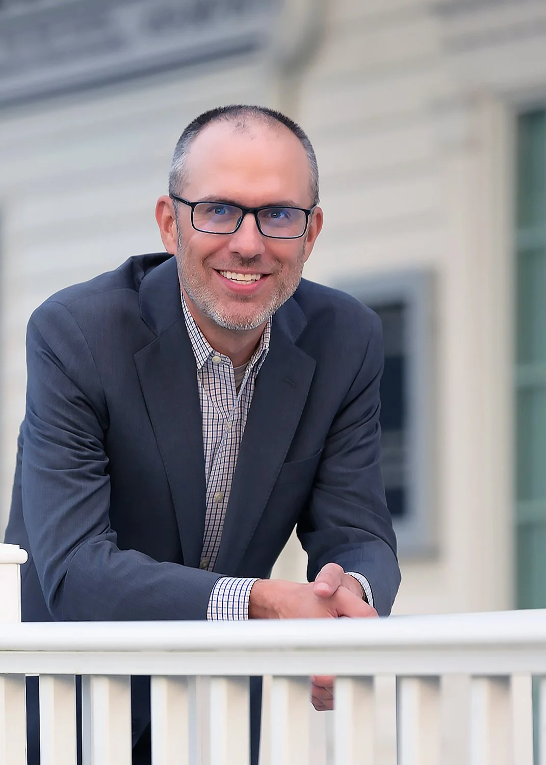 A smiling man with glasses, wearing a dark suit jacket and a checkered shirt, leaning on a white railing in front of a residential house.
