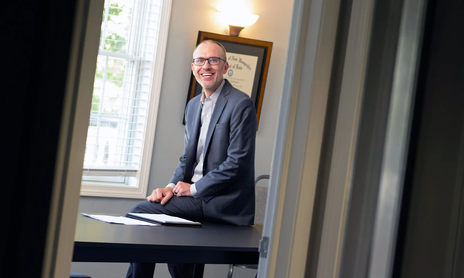 A man in a suit and glasses sitting on a table, smiling through a doorway in an office setting.