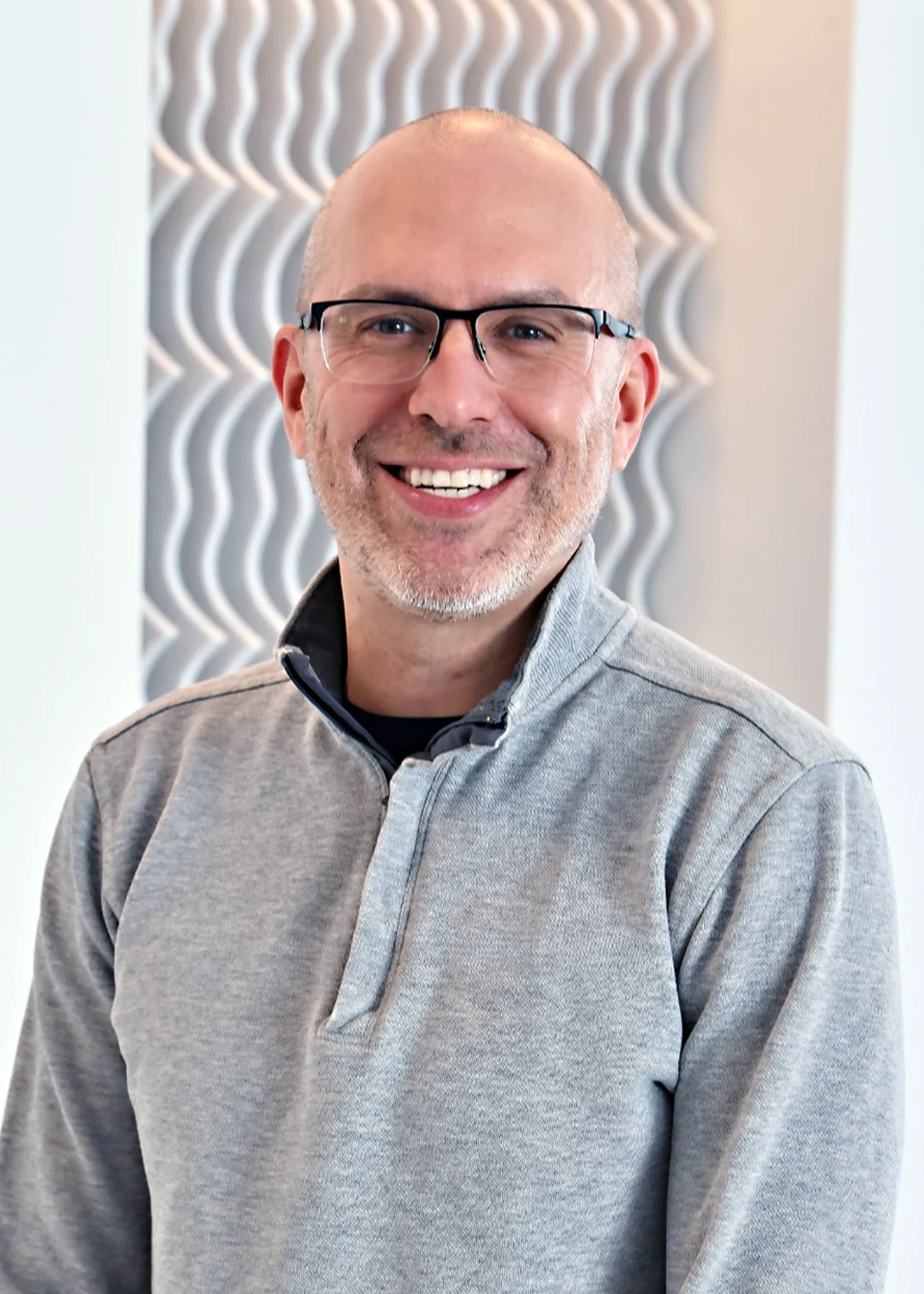 A smiling man with glasses, a beard, and a shaved head wearing a light gray zip-up sweater, standing indoors against a textured white wall.